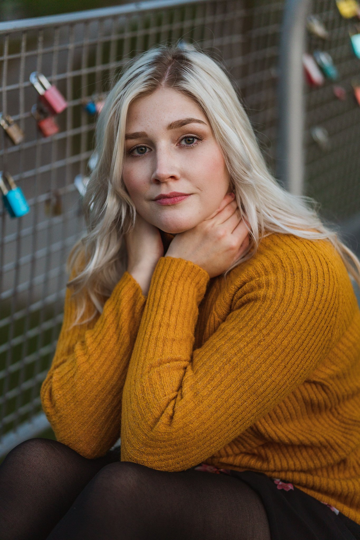 Junge Frau mit blonden, gewellten Haaren in einem braungelben Pullover, sitzend vor einem Metallzaun mit Vorhängeschlössern, schaut in die Kamera.