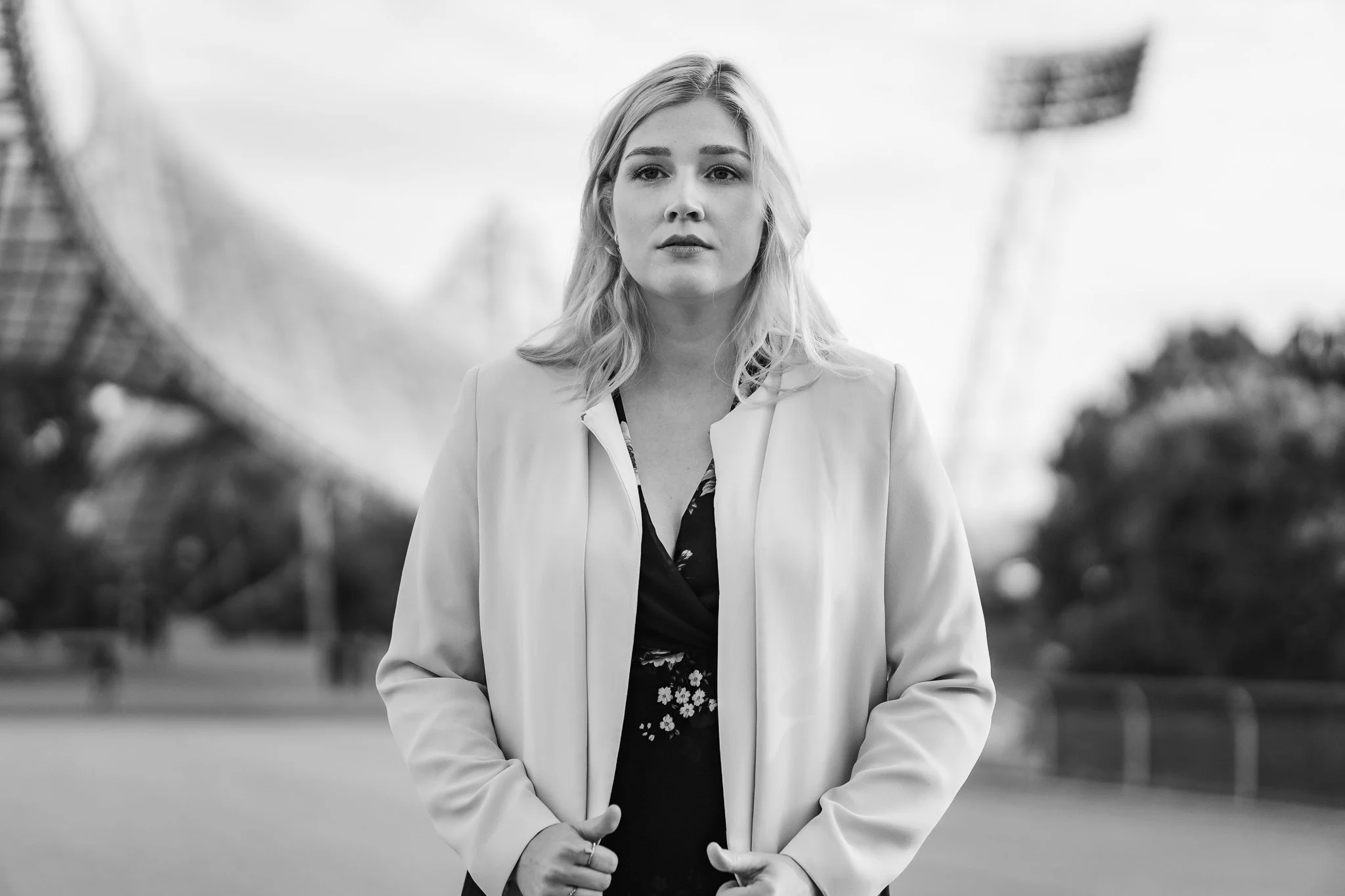 Junge Frau mit blonden Haaren in einem Park, schwarzen Oberteil mit Blumenmuster, hell-helle Jacke, nachdenklicher Ausdruck, im Schwarz-Weiß-Foto.