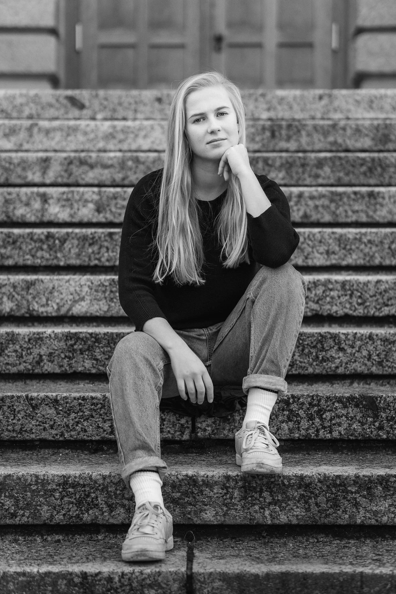 Junge Frau mit langen Haaren sitzt nachdenklich auf Steinstufen vor einem Holztor in Schwarz-Weiß