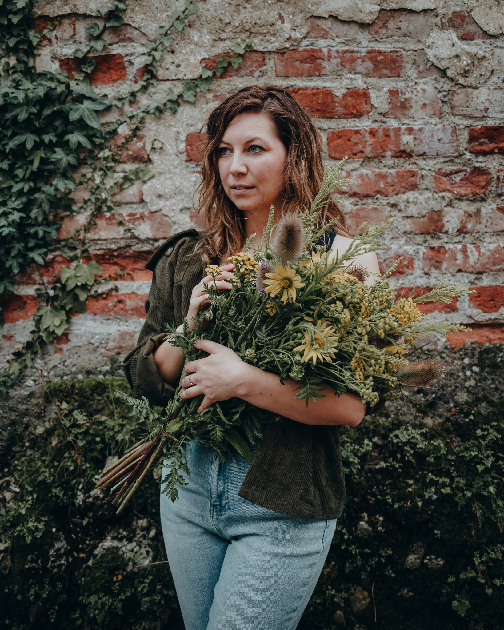 Junge Frau mit braunen, lockigen Haaren hält einen großen Blumenstrauß vor einem Backsteinmauer. Sie trägt eine grüne Jacke und blaue Jeans.