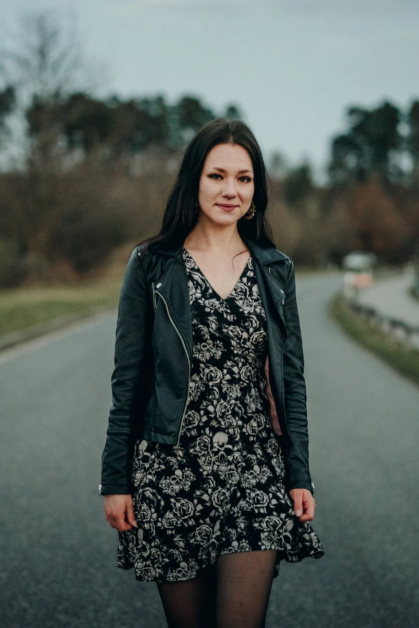 Eine junge Frau steht auf einer papieren Straße im Freien, trägt ein schwarzes Lederjacke und ein schwarzes Kleid mit weißen Rosenmuster, hat dunkle Haare, leichte Make-up und blickt in die Kamera.