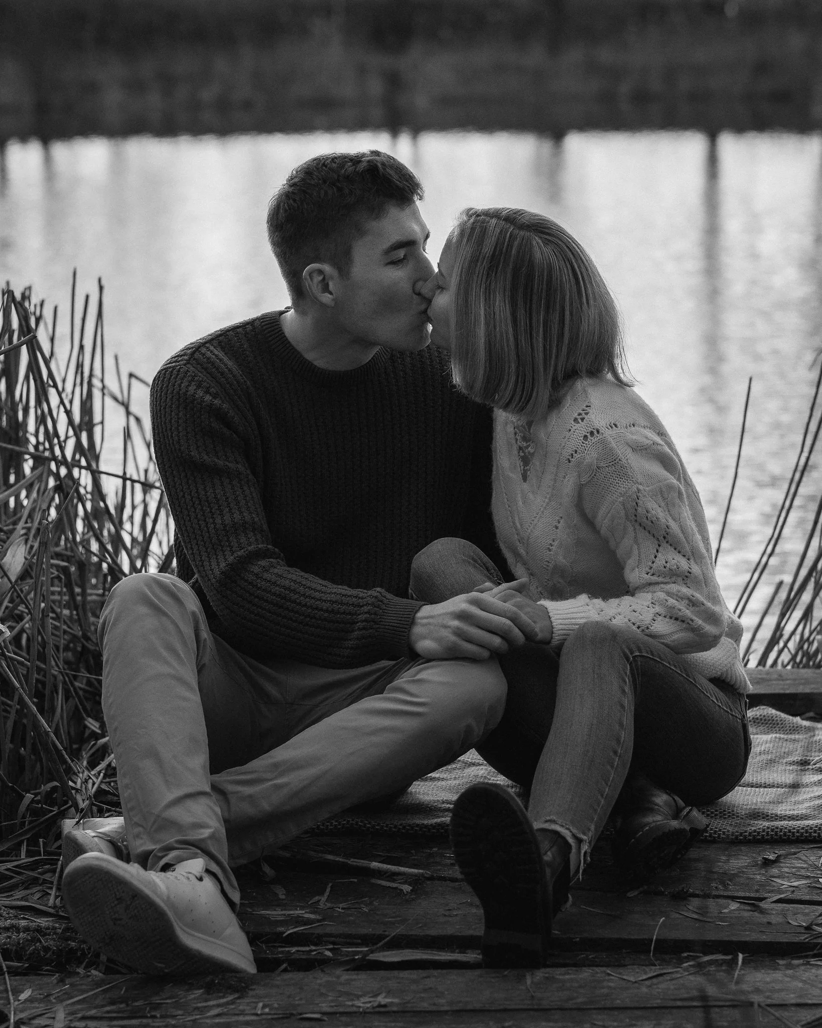 Ein junges Paar sitzt am Wasser und küsst sich leidenschaftlich auf einer Holzplattform, umgeben von Schilf, in schwarz-weiß.