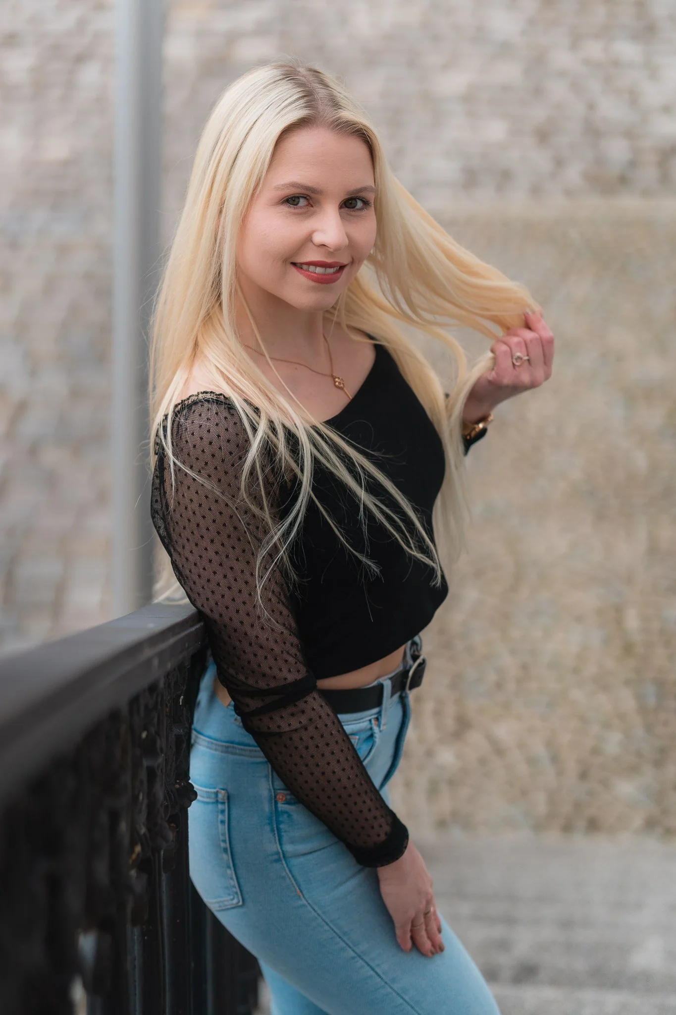 Junge blonde Frau in schwarzen Top mit Mesh-Ärmeln und hellblauen Jeans steht an einem Geländer und lächelt.