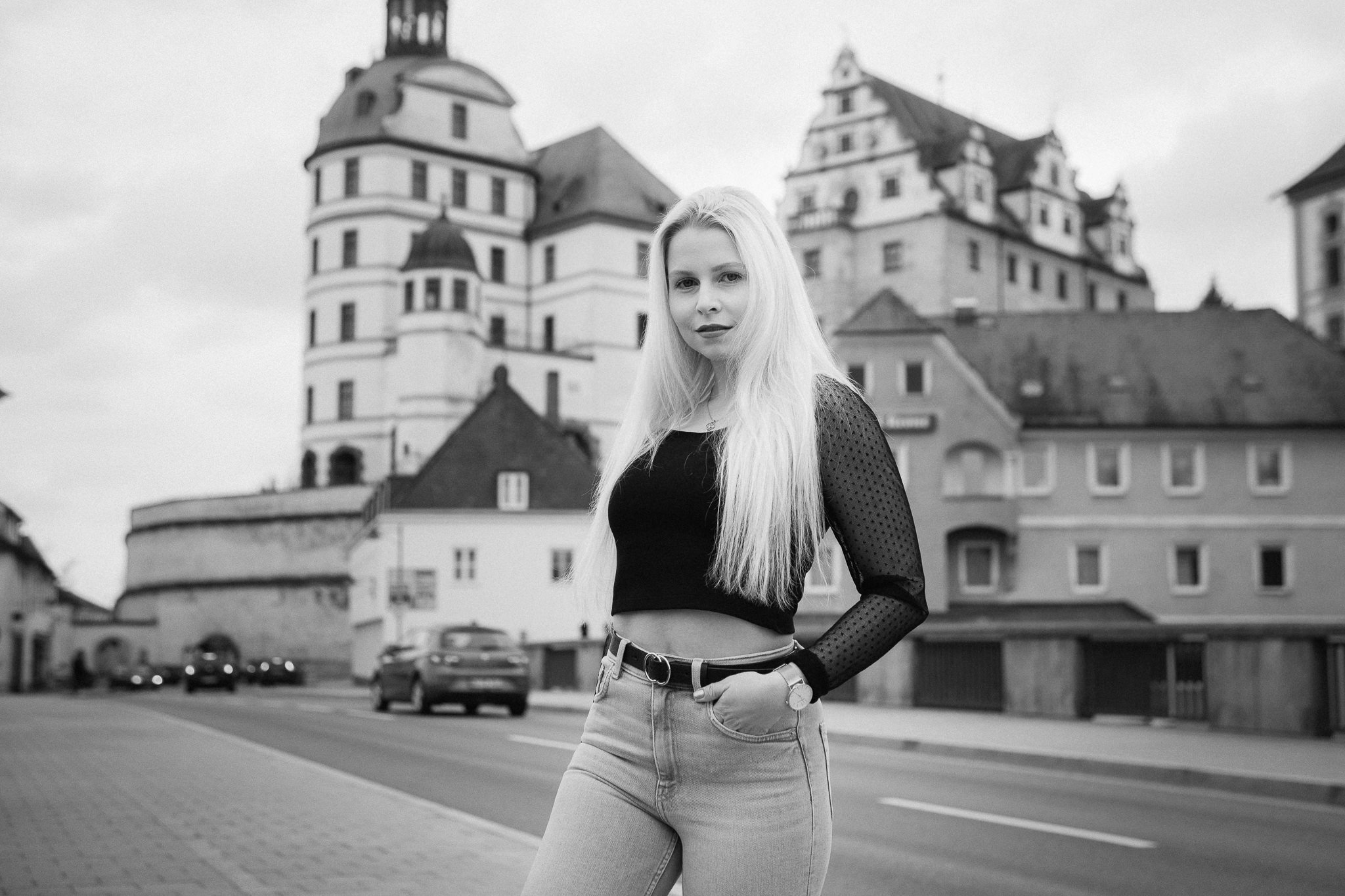 Junge Frau mit langen hellen Haaren trägt schwarzen Crop-Top mit transparenten Ärmeln und Jeans, steht auf einer Straße vor einem historischen Schloss.