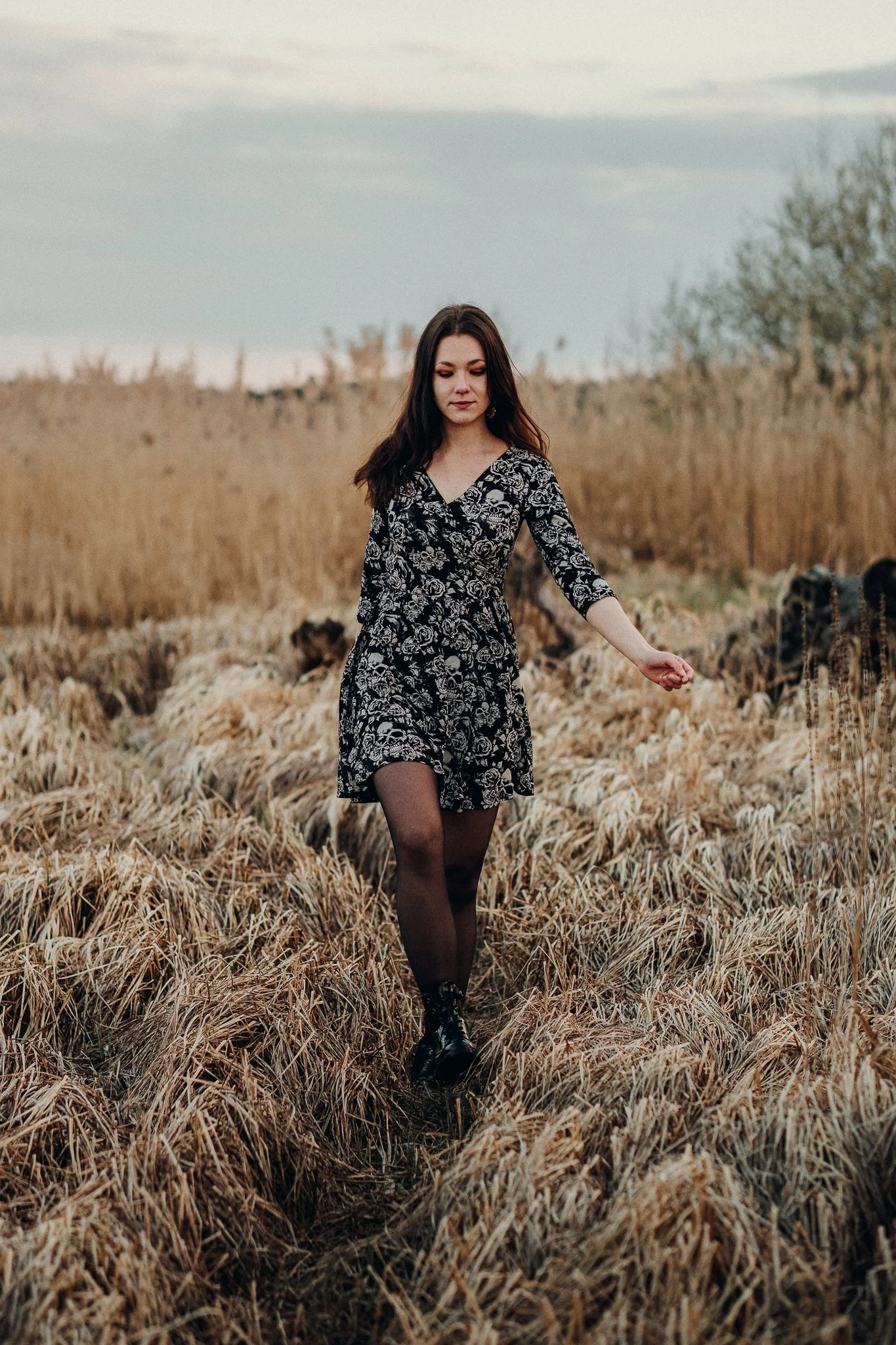 Eine junge Frau in einem schwarzen Kleid mit weißen Mustern, trägt schwarze Stiefel und läuft durch ein Feld mit trockenen Gräsern, im Hintergrund sind Bäume und ein bewölkter Himmel.
