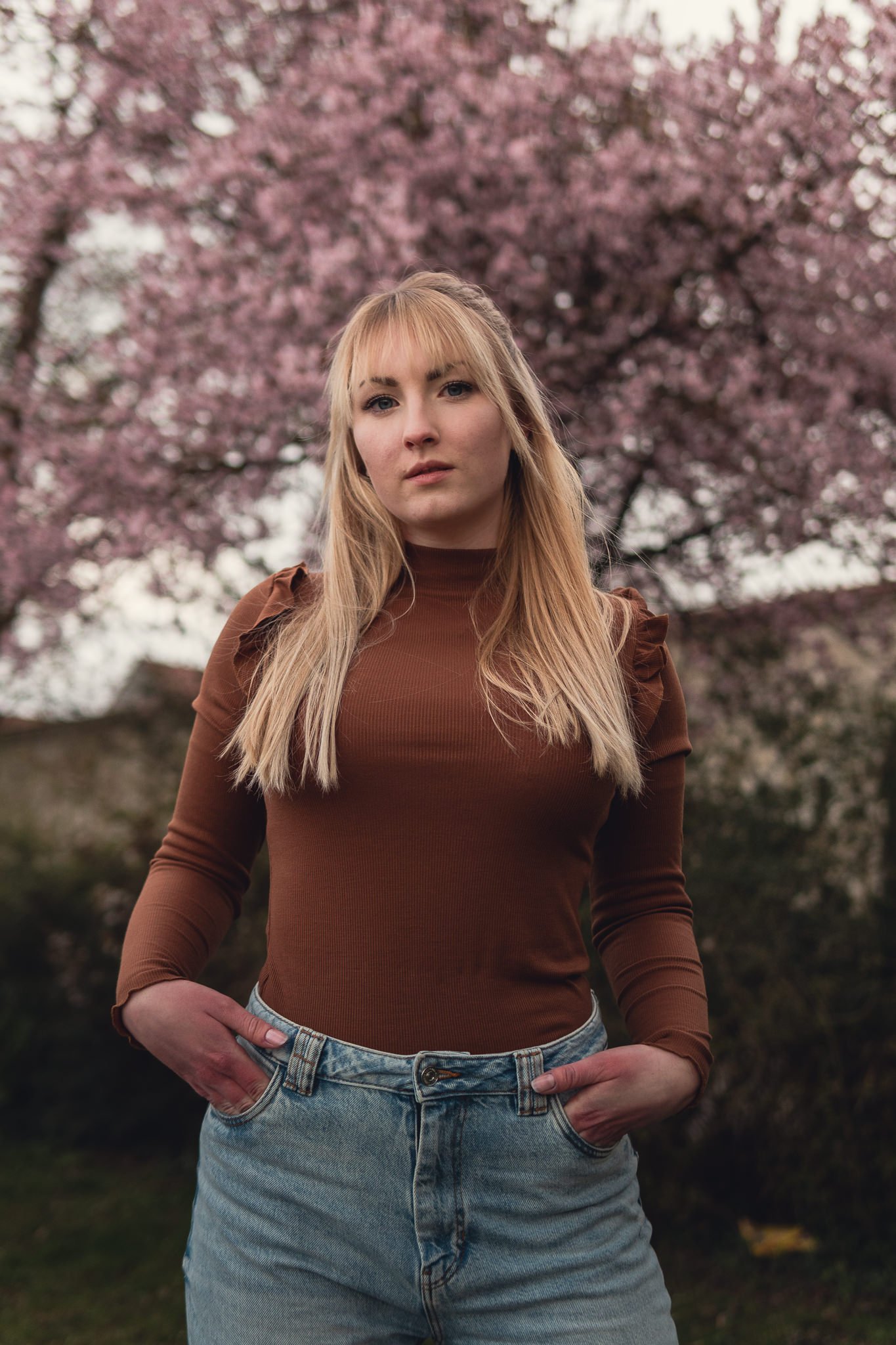 Junge Frau steht vor blühender Kirschbaum im Park, trägt brauen Pullover und Jeans, Blick in die Kamera.