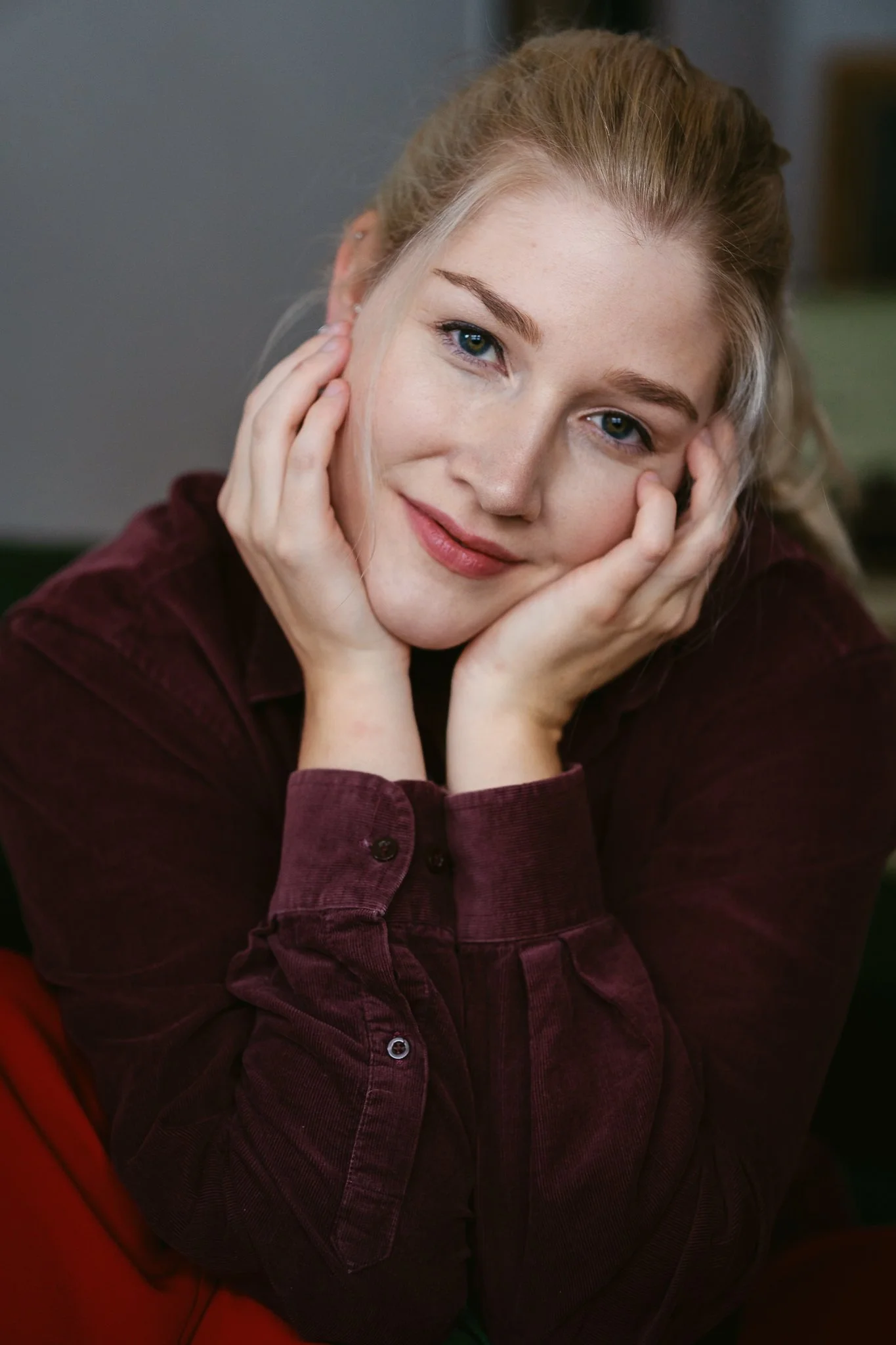 Nahaufnahme einer jungen Frau mit blonden Haaren, die lächelt und ihre Hände an ihrem Gesicht hält, trägt einen dunkel rotn Anzug.