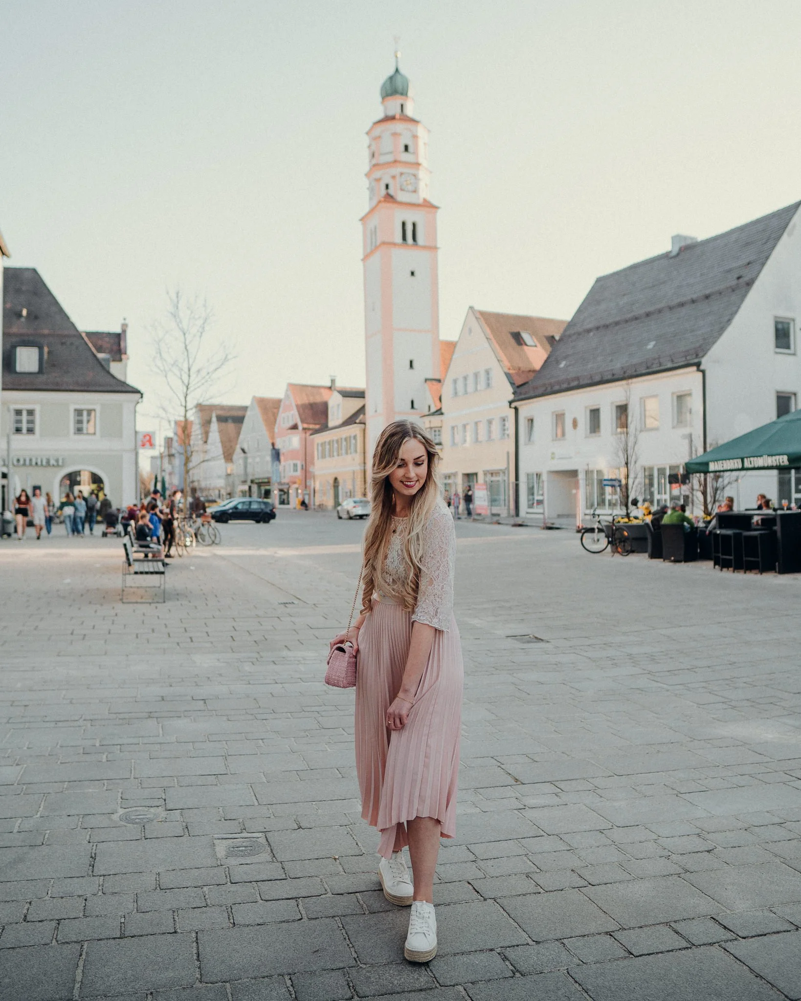 Junge Frau geht in einer Stadt mit historischen Gebäuden und einem Kirchturm spazieren, trägt ein langes, hellrosa Kleid und weiße Turnschuhe.