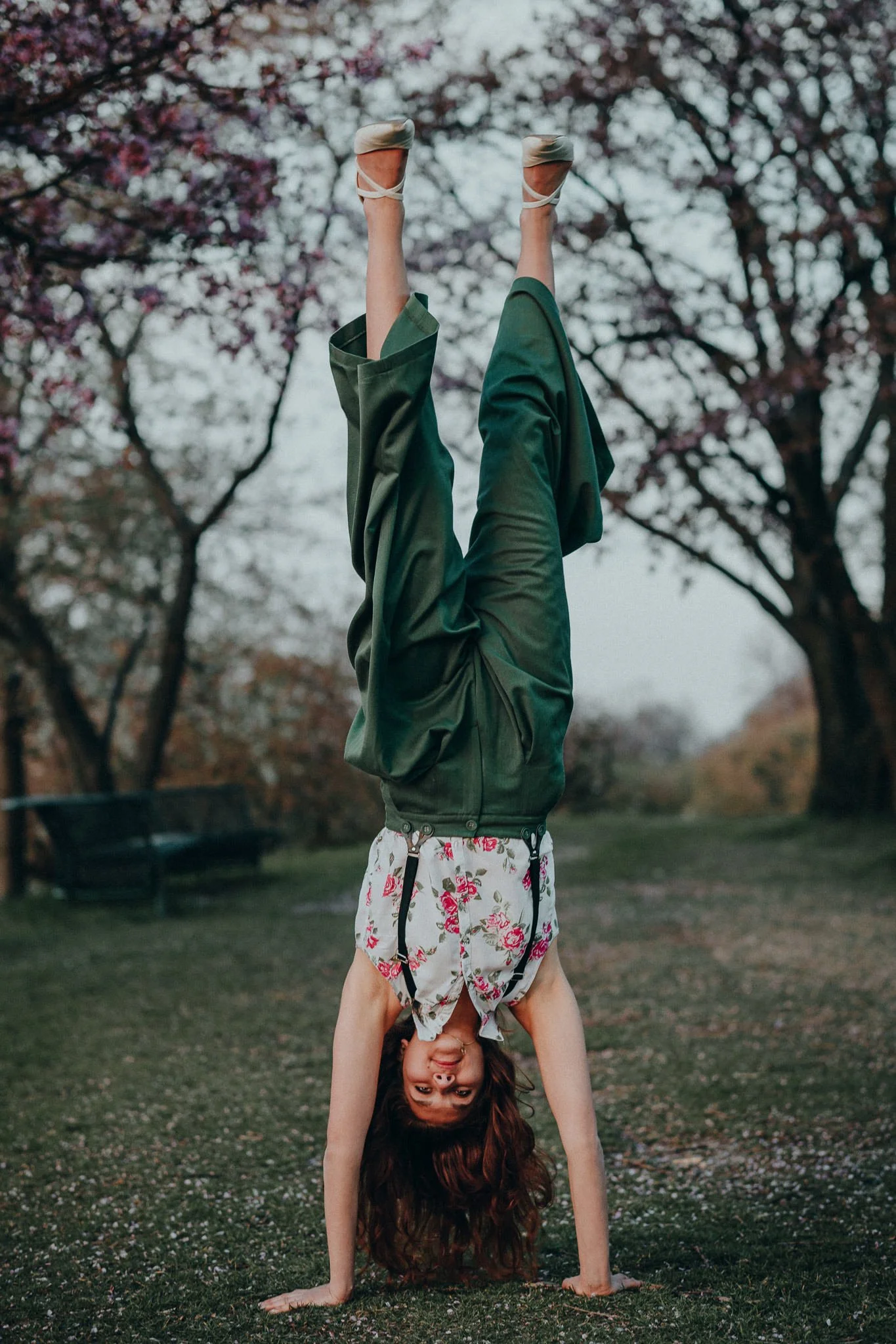 Frau macht Handstand auf Rasen in Park, Bäume mit Blüten im Hintergrund, trägt Shirt mit Blumenmuster und grüne Hosen.