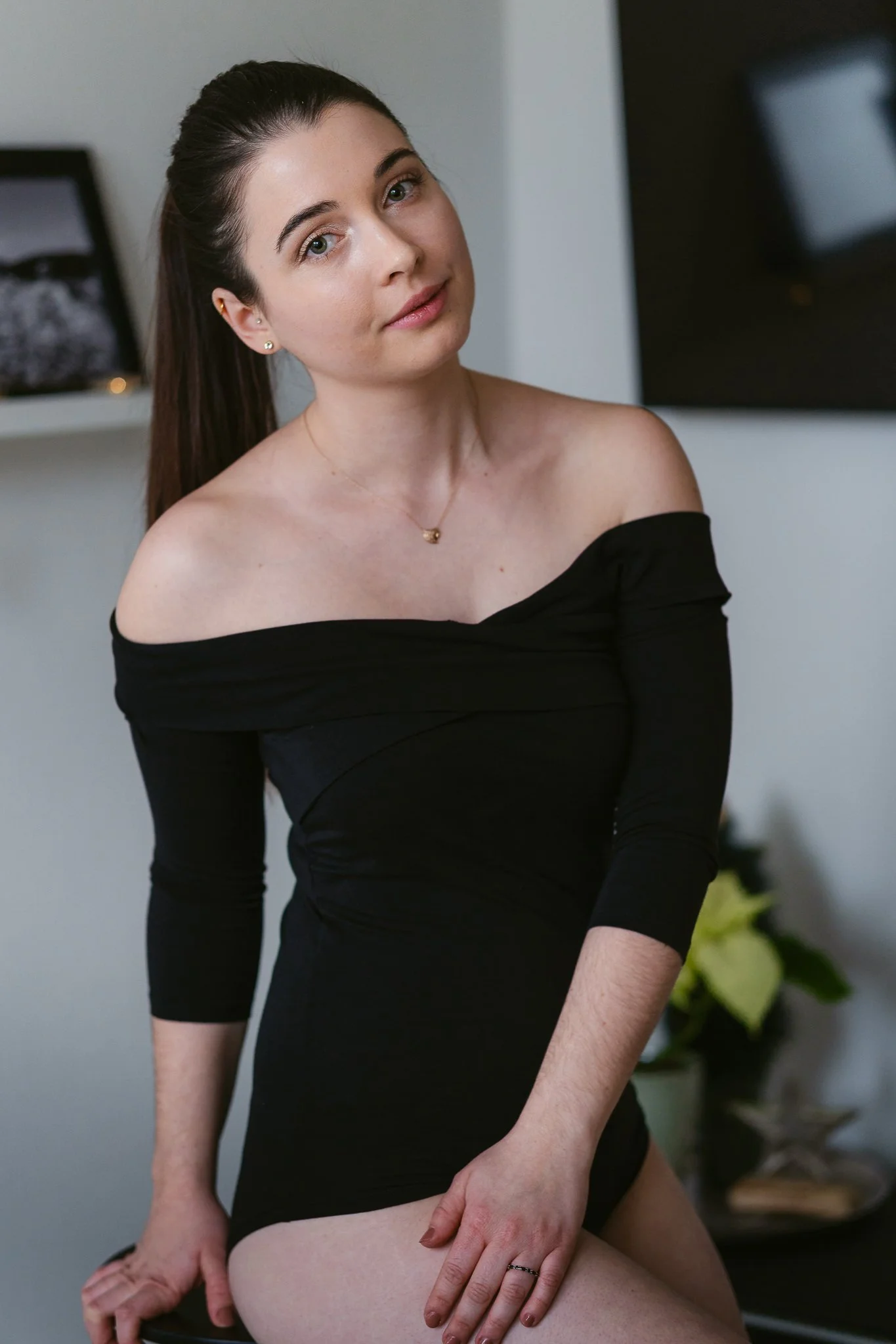 Junge Frau mit dunklem Haar, trug ein schwarzes, off-shoulder Kleid, sitzt in einem modernen Raum, Blick in die Kamera gerichtet.