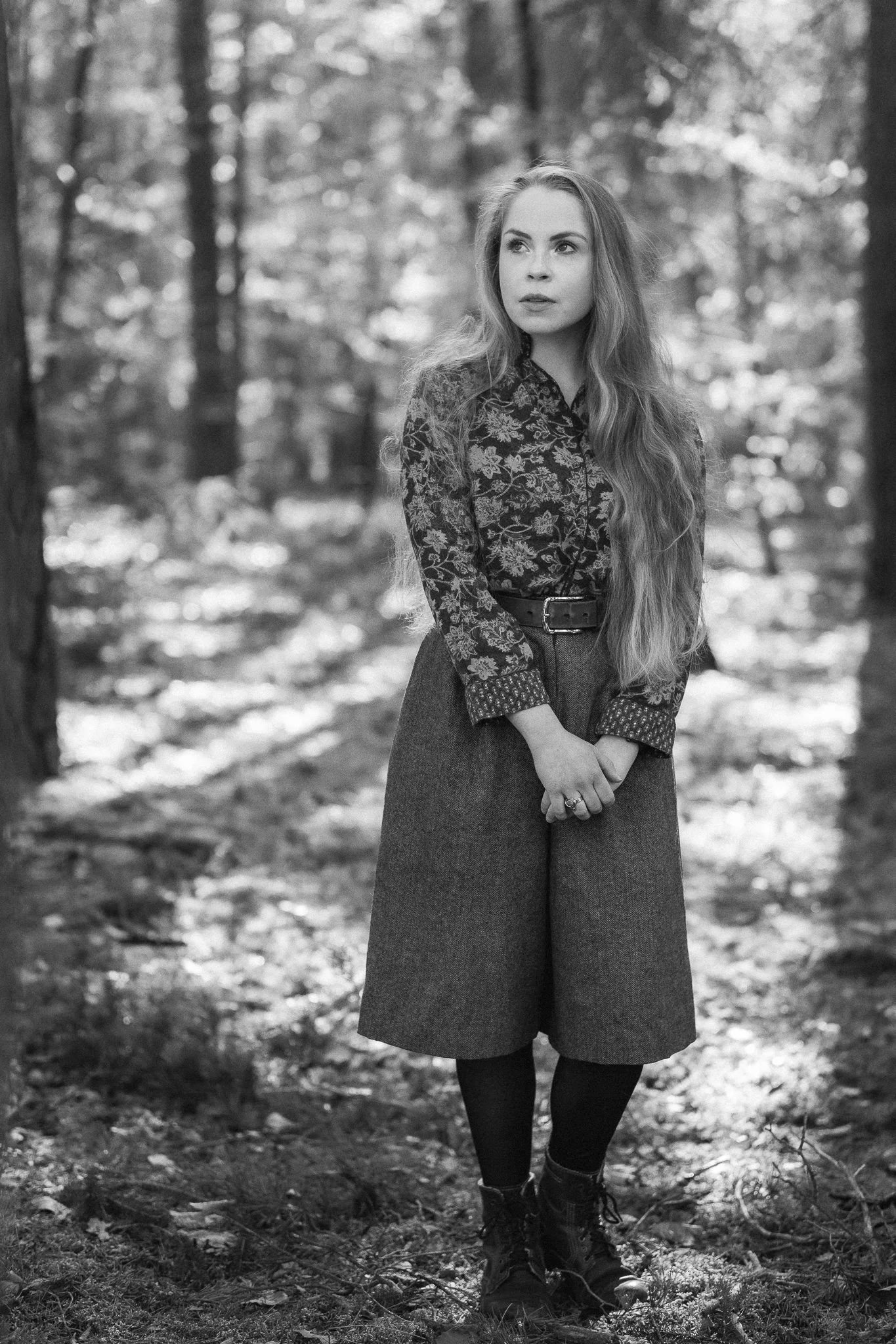 Schwarz-Weiß-Foto einer jungen Frau mit langen Haaren in einem Wald, trägt ein langärmeliges, floral gemustertes Hemd, eine hoch taillierte Hose und Schuhe, steht nachdenklich zwischen Bäumen.