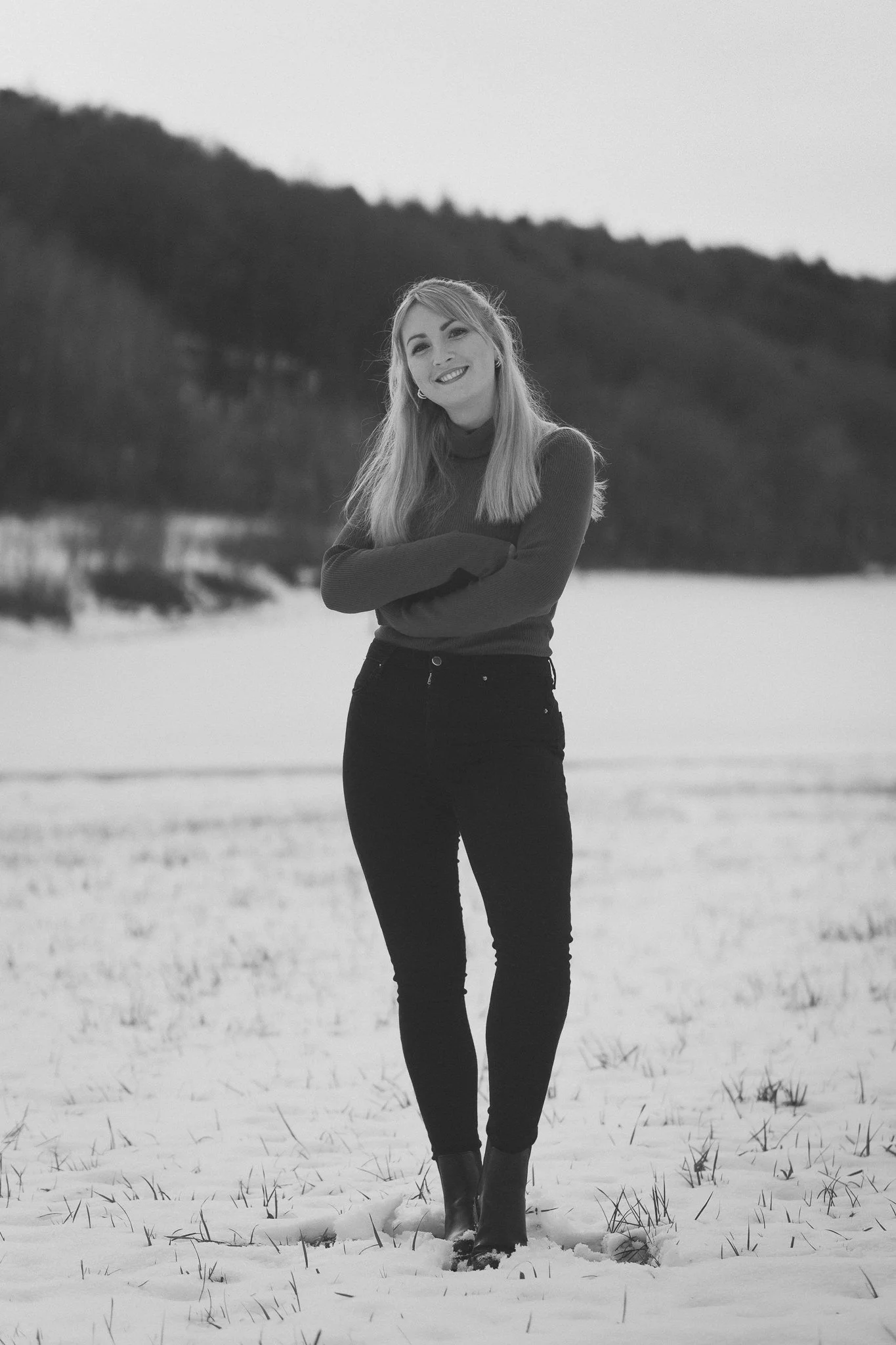 Schwarz-Weiß-Foto einer lachenden jungen Frau mit langen Haaren, armkreuzend, in einer Schneelandschaft mit Hügeln im Hintergrund.