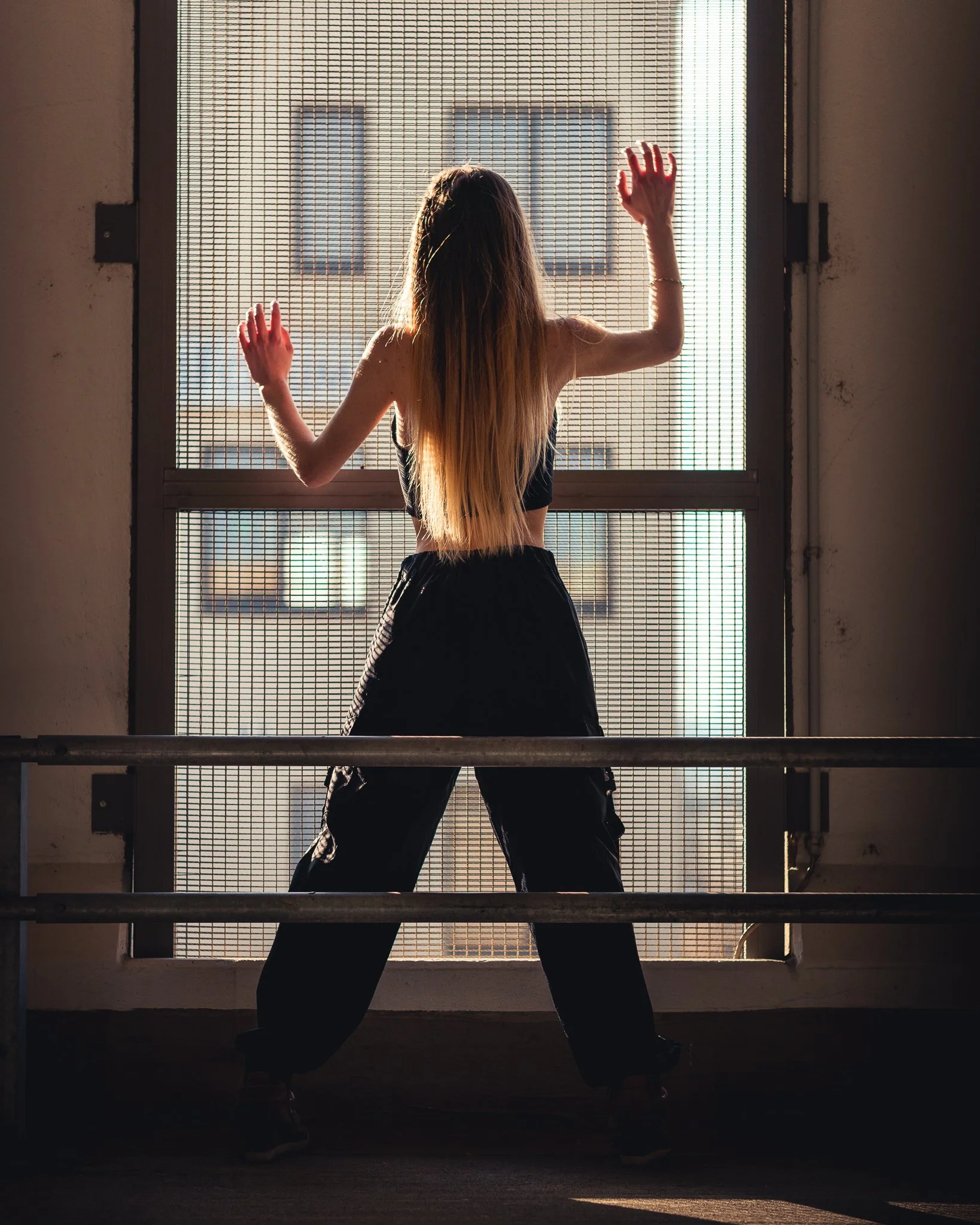 Frau mit langen Haaren steht vor einem Gitterfenster in einem Raum, Blick nach draußen, Sonnenlicht erleuchtet den Raum.