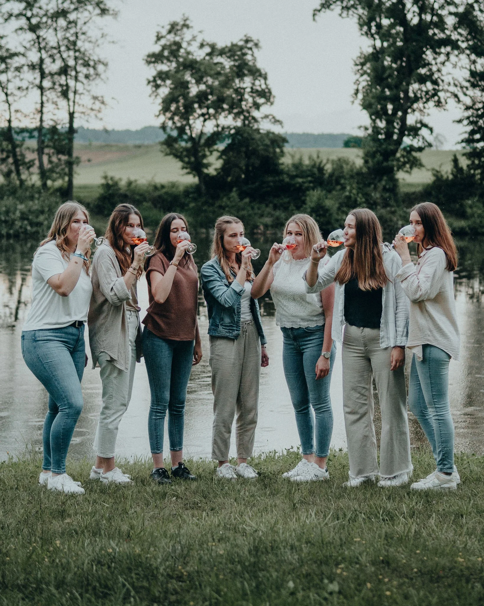 Eine Gruppe von acht Frauen steht am Ufer eines Sees und trinkt Bier aus Gläsern, umgeben von Bäumen und grünen Wiesen.