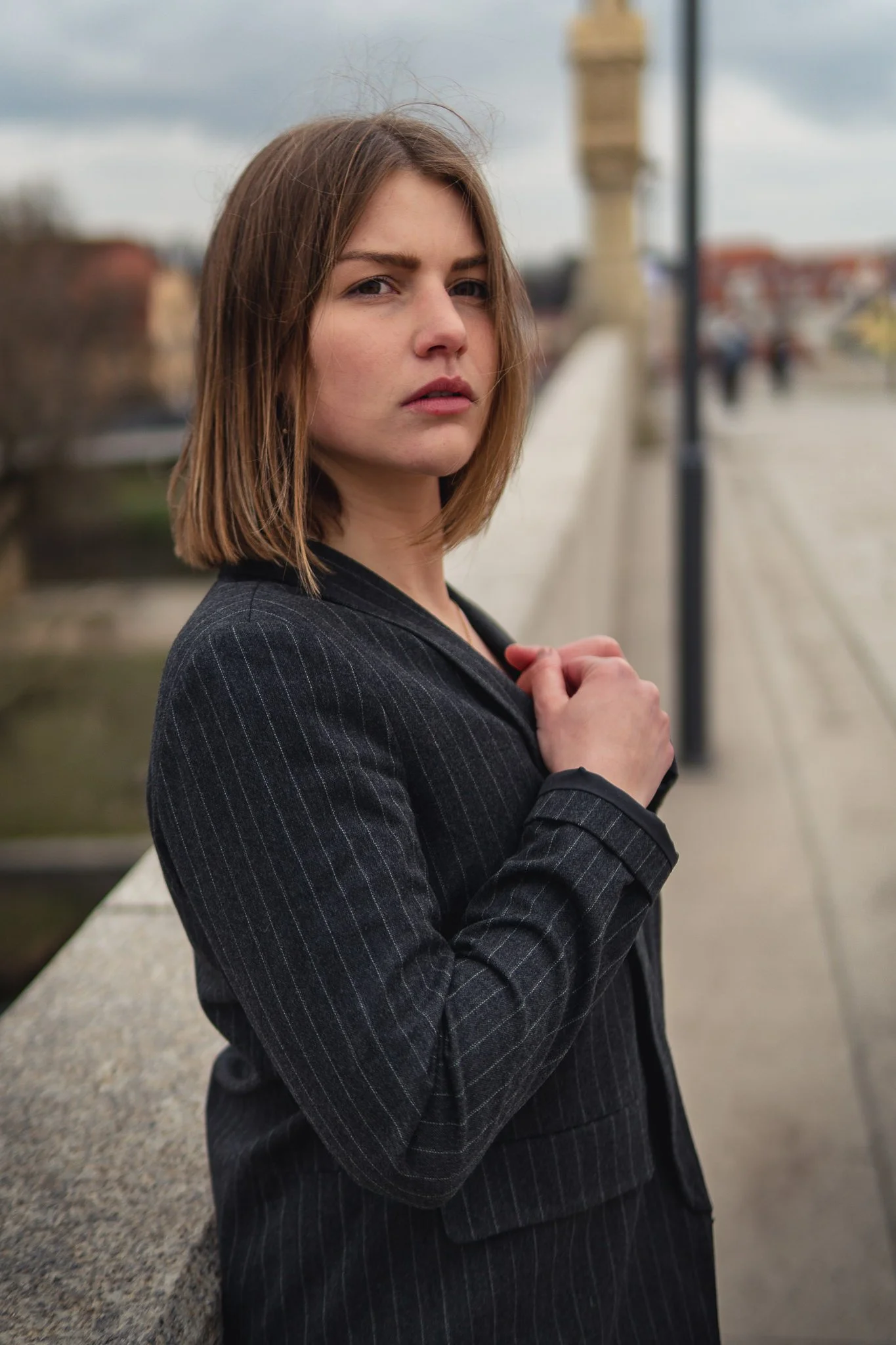 Junge Frau in dunklem Anzug auf Brücke, Blick zur Kamera, im Hintergrund Wolkenverlauf.