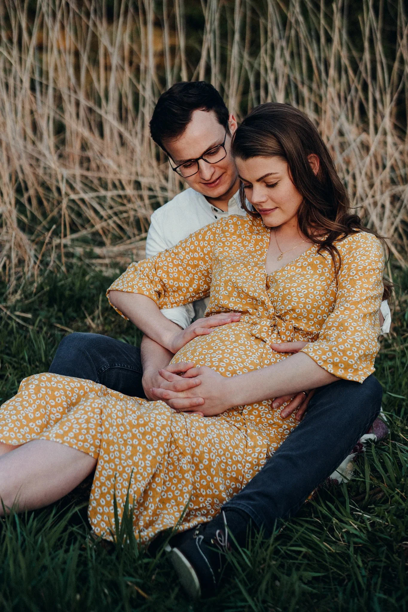 Ein Paar sitzt auf dem Boden in einem Feld, die schwangere Frau trägt ein gelbes Kleid mit weißen Mustern, der Mann umarmt sie liebevoll.