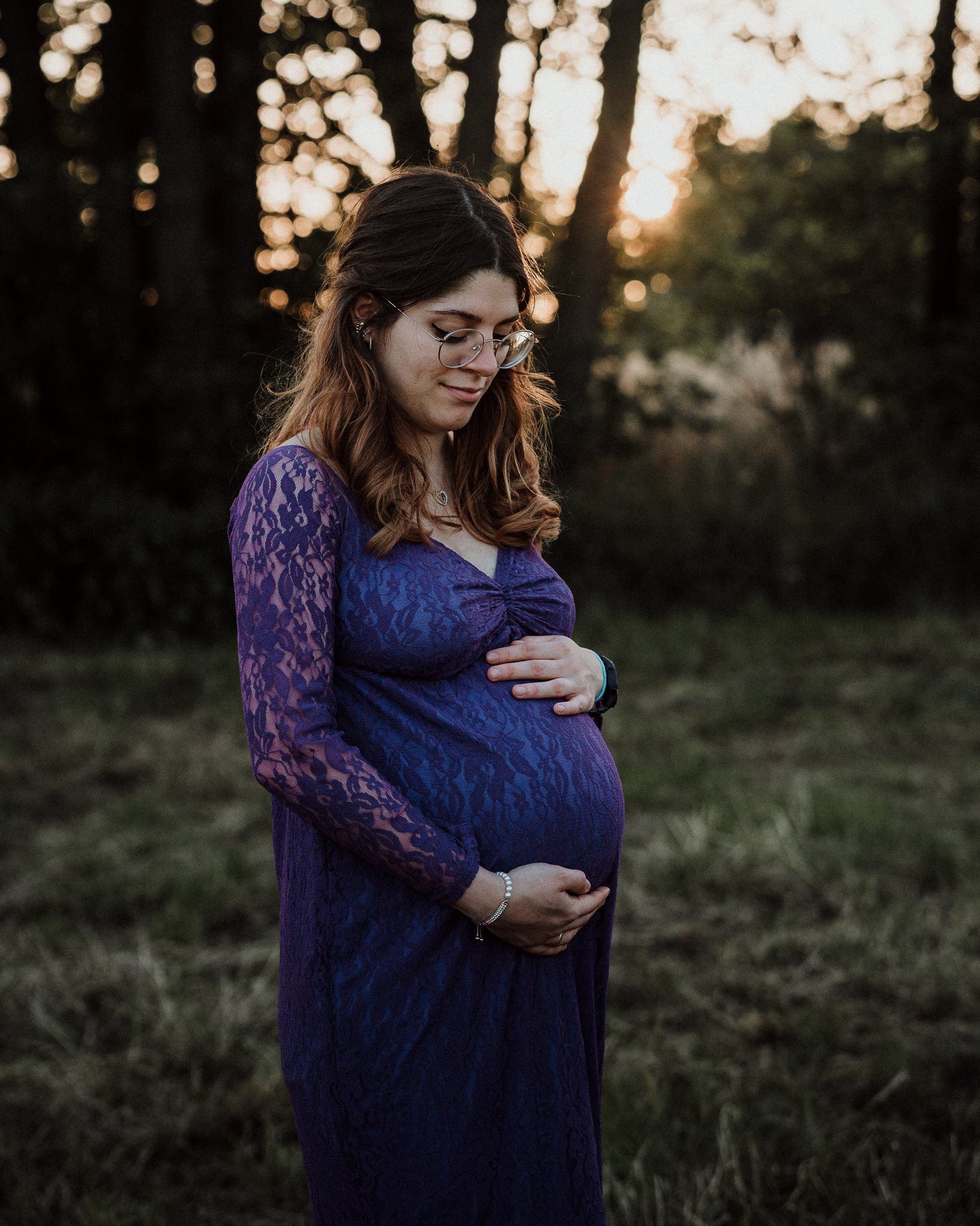 Schwangere Frau in einer lila Spitzenkleidung steht im Freien bei Sonnenuntergang, hält ihren Babybauch und schaut ihn liebevoll an.
