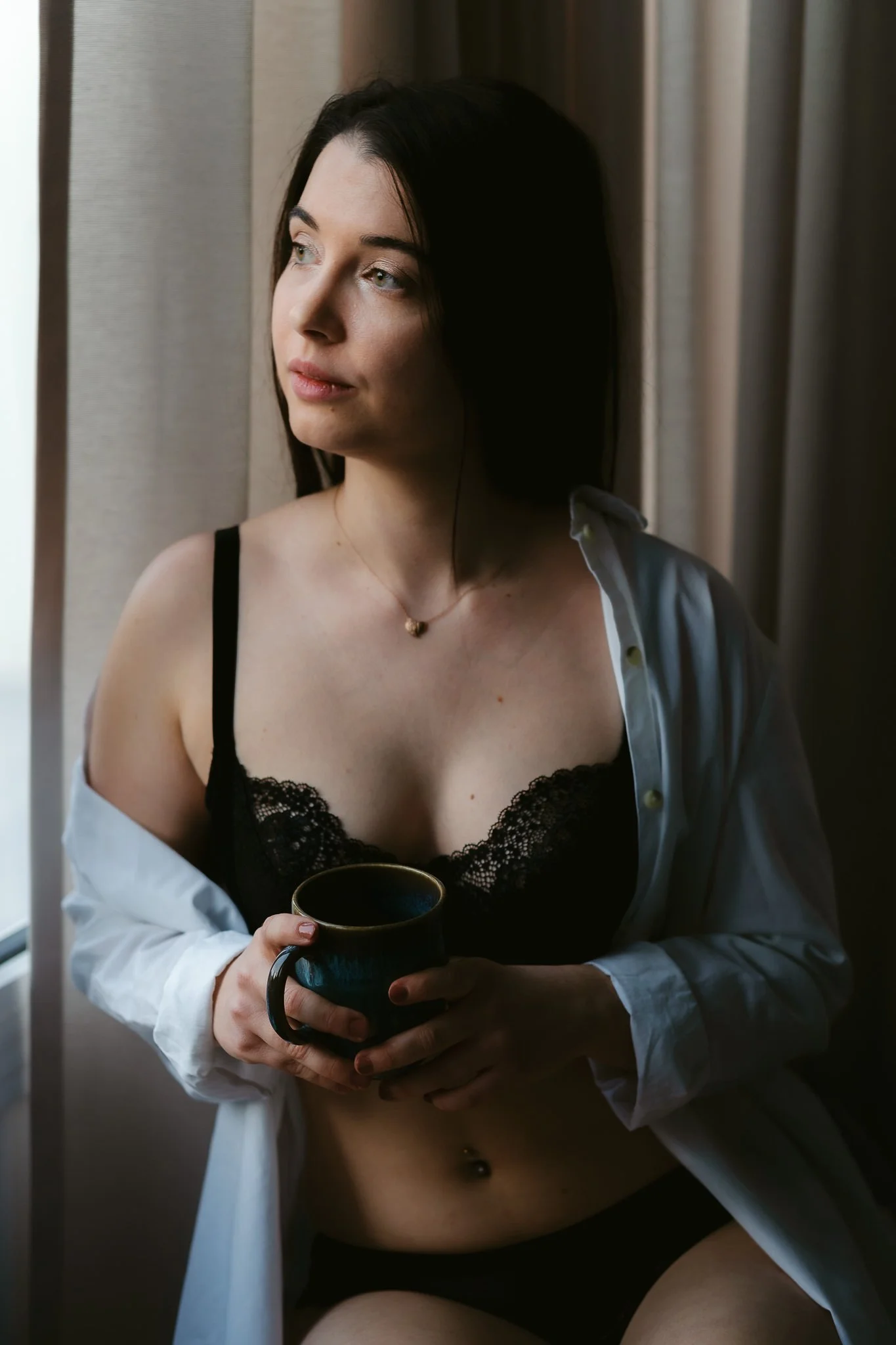 Junge Frau mit dunklem Haar, die in einem schwarzen Spitzen-BH und ungeknöpfter weißen Hemd sitzt und eine Tasse hält, während sie aus dem Fenster blickt.