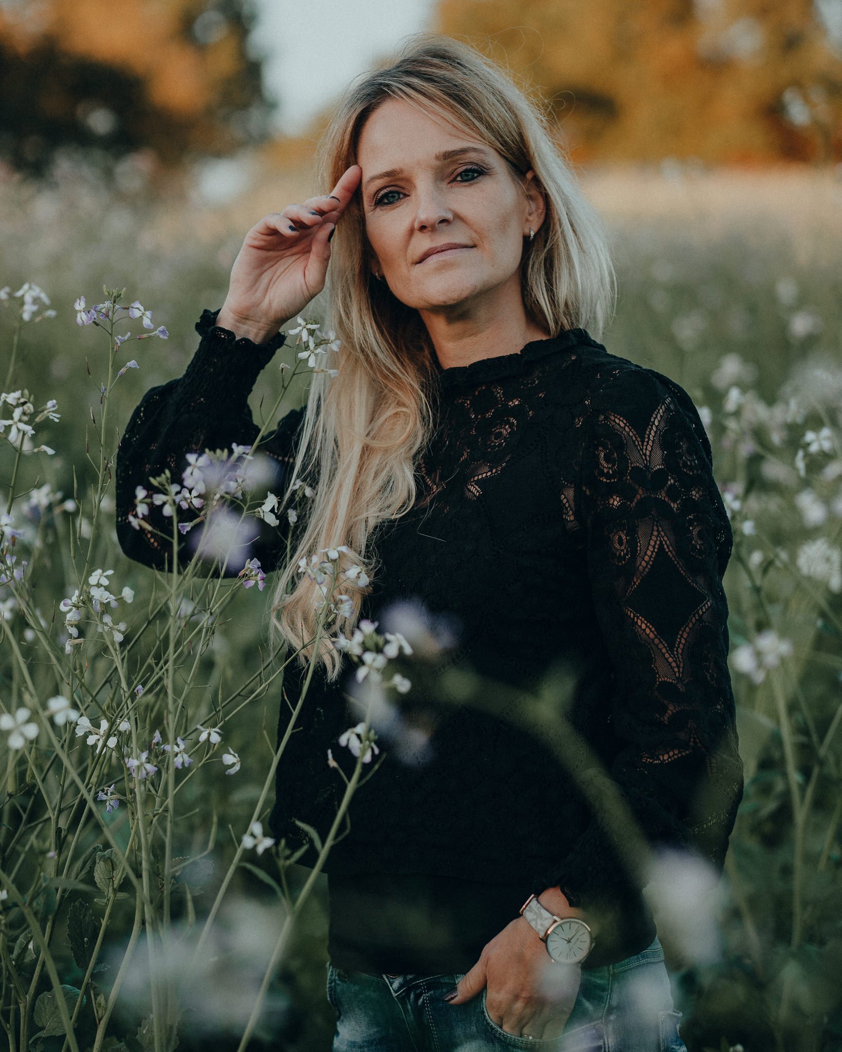 Eine Frau mit langem blondem Haar in einer schwarzen, aus Spitze bestehenden Bluse steht inmitten von blühenden Pflanzen in einem Feld, während sie nachdenklich in die Ferne schaut.