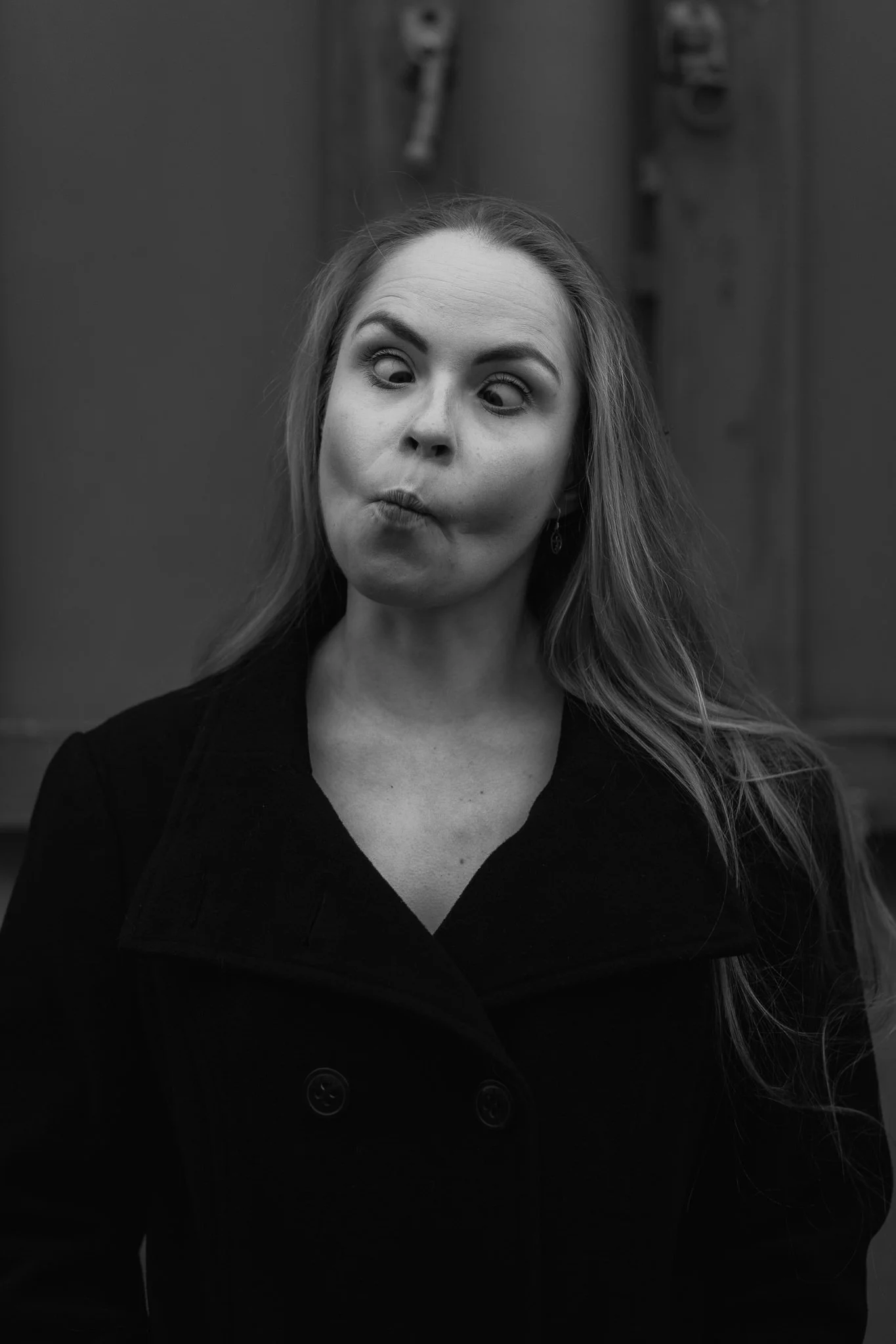 Schwarz-Weiß-Foto einer Frau, die eine lustige Grimasse macht, mit hochgezogenen Augenbrauen und leicht geschürzten Lippen, vor einem dunklen Hintergrund.