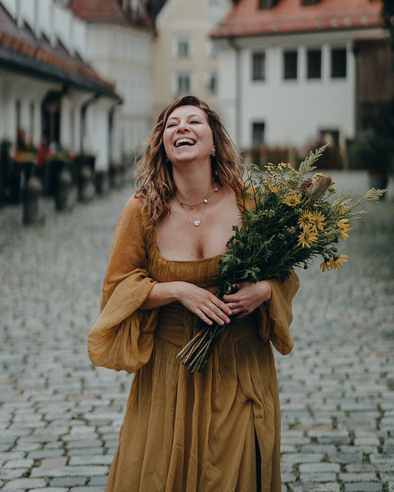 Eine lachende Frau in einem gelben Kleid hält einen Blumenstrauß auf einer gepflasterten Straße mit historischen Gebäuden im Hintergrund.