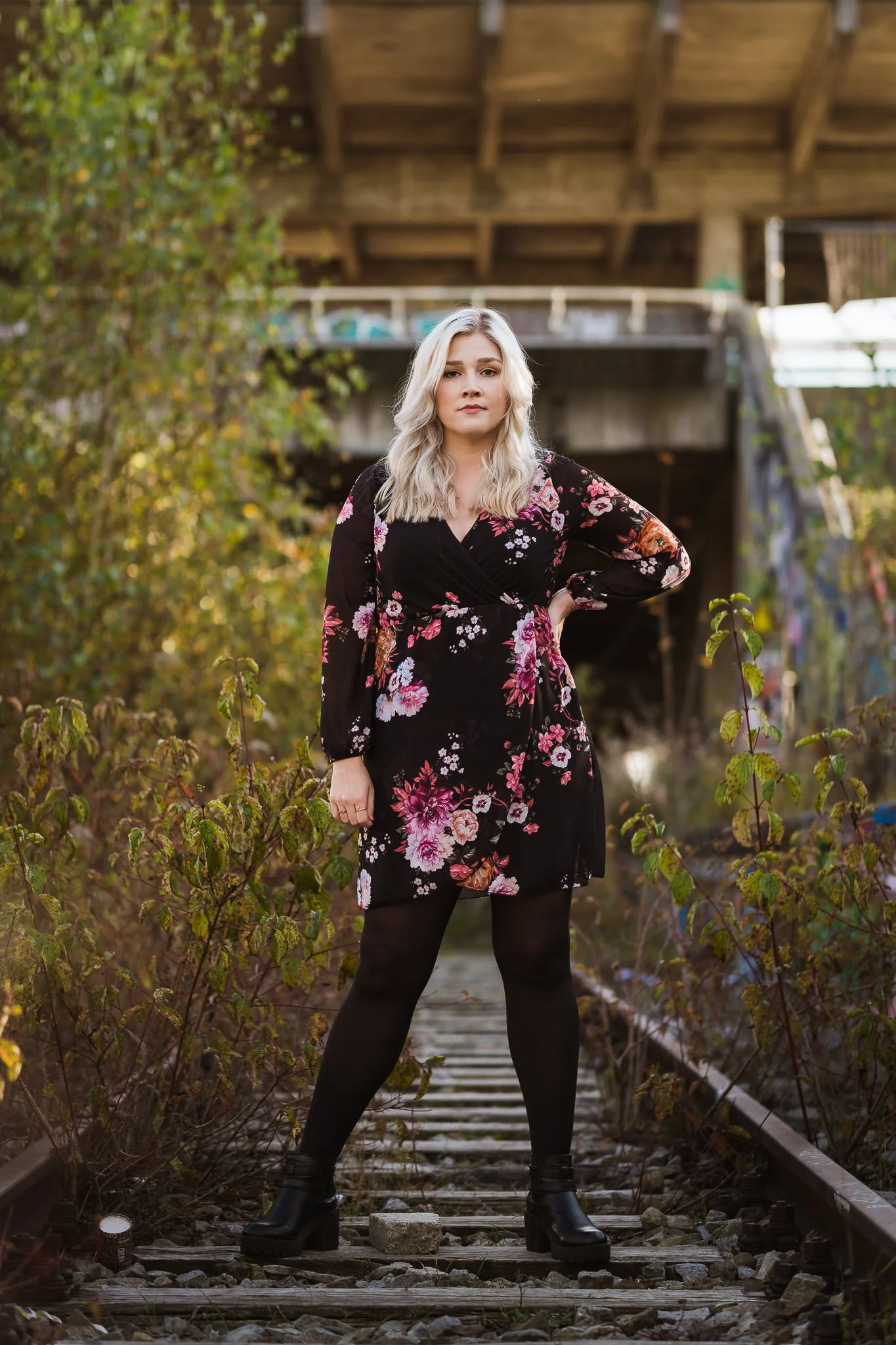 Junge Frau im schwarzen, floral gemusterten Kleid und schwarzen Stiefeln steht auf einen alten Eisenbahn-Schiene, umgeben von Büschen, im Freien, mit einem Holzgang im Hintergrund.