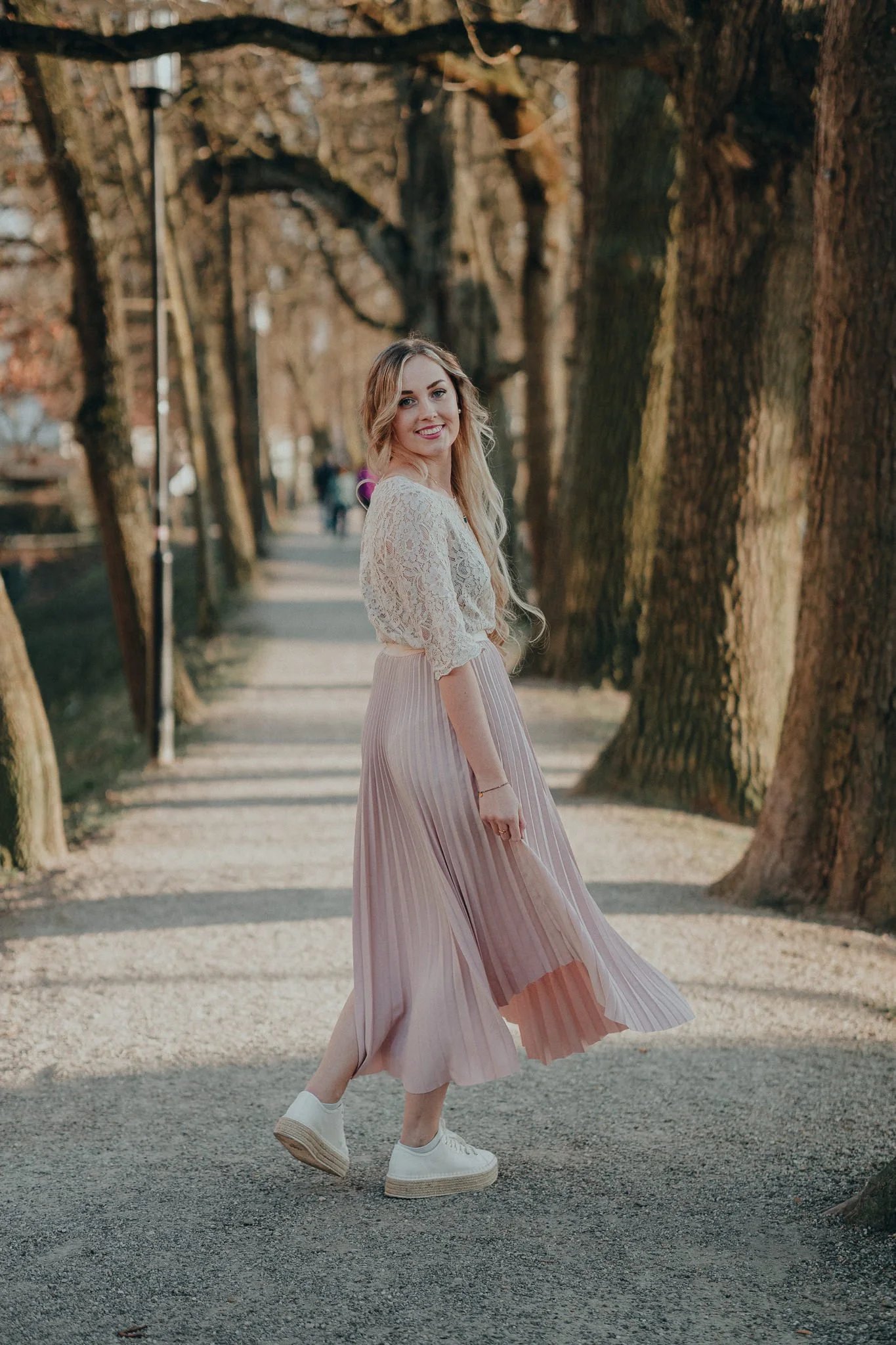 Eine lächelnde junge Frau in einem weißen Spitzen-Oberteil und einem rosa, plissierten Rock mit weißen Sneakers, die auf einem Baumweg in einem Park im Herbst steht.