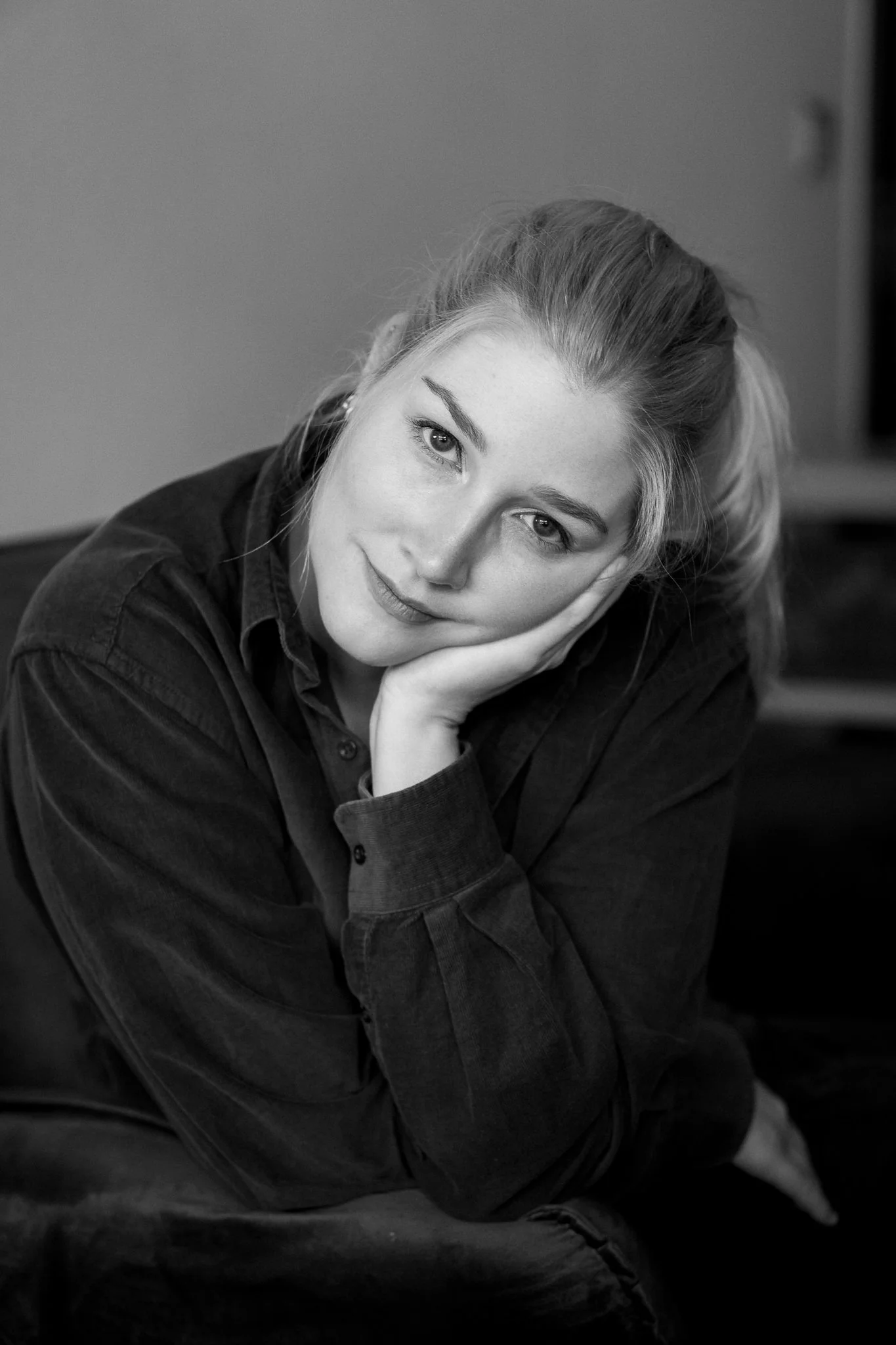 Ein schwarz-weiß Foto einer jungen Frau mit hellen Haaren, die ihre Wange mit ihrer Hand stützt und direkt in die Kamera schaut.