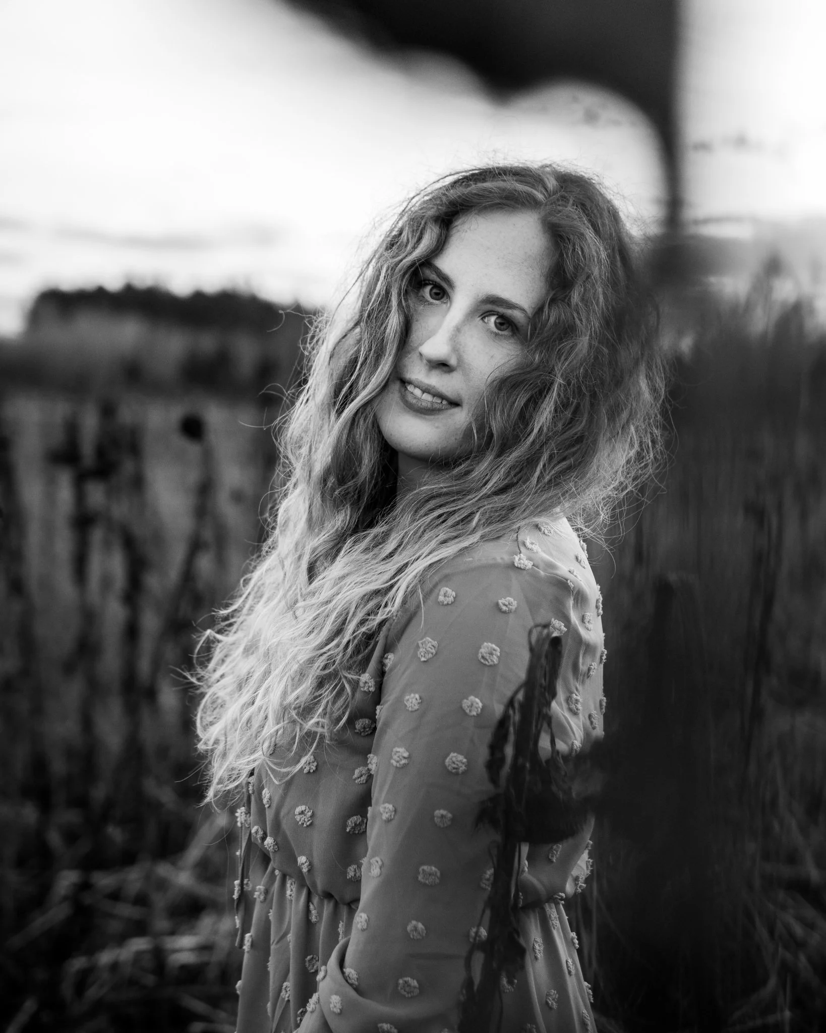 Schwarz-Weiß-Fotografie einer jungen Frau mit langen, lockigen Haaren, die in einem Feld steht, mit einem sanften Blick in die Kamera.