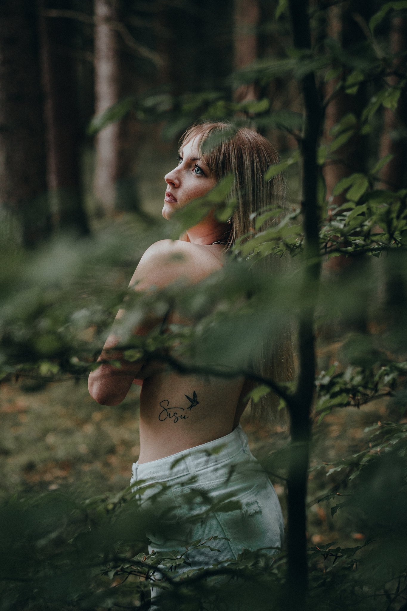 Eine junge Frau mit braunen Haaren steht in einem Wald, umgeben von Blättern. Sie trägt weiße Hosen und hat ein Tattoo mit dem Wort "Sisu" und einer Vogelfeder auf ihrer linken Seite. Sie blickt nach oben, ihre Arme sind verschränkt vor der Brust.