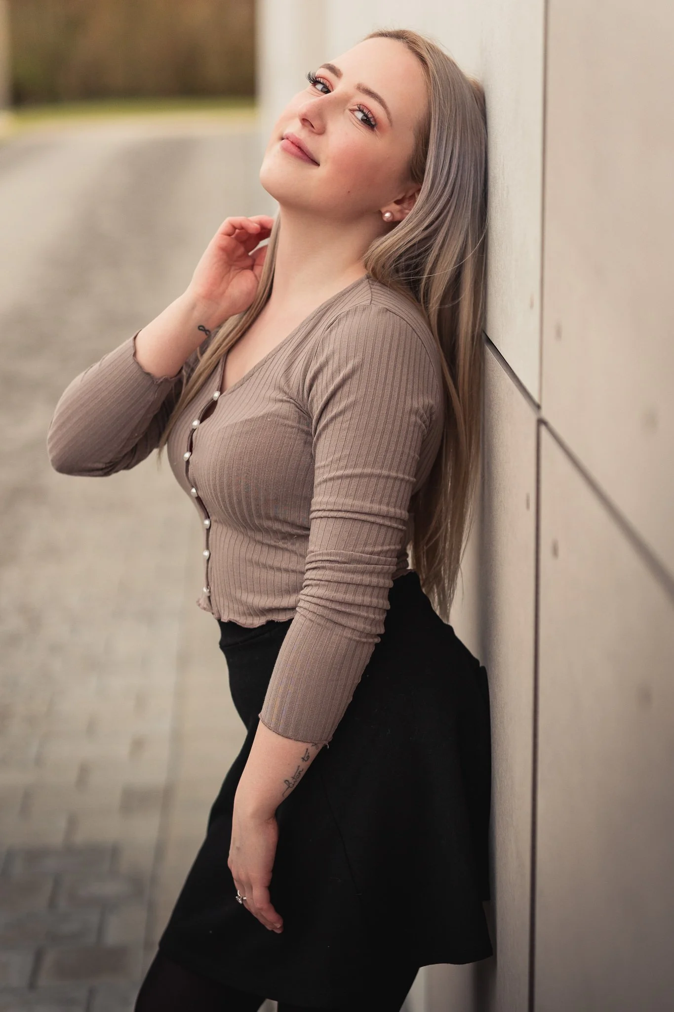 Junge Frau lehnt an einer Wand im Freien, trägt ein braunes, langärmeliges, gestreiftes Oberteil und eine schwarze Skirt, hat lange blonde Haare, lächelt leicht, im Hintergrund Bäume und einen weichen, unscharfen Park.