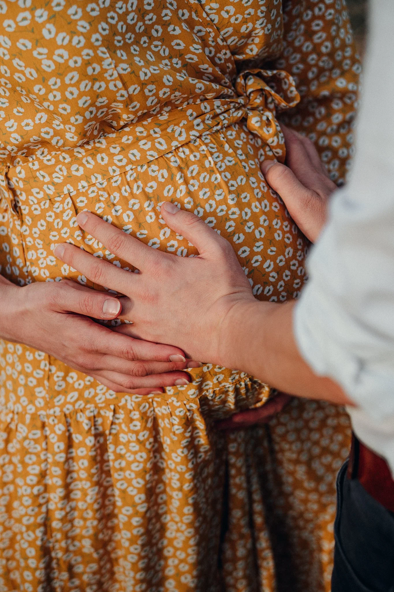 Eine schwangere Person trägt ein orangefarbenes Kleid mit weißen Blumenmustern. Jemand anderes berührt ihren Bauch und legt die Hand darauf.