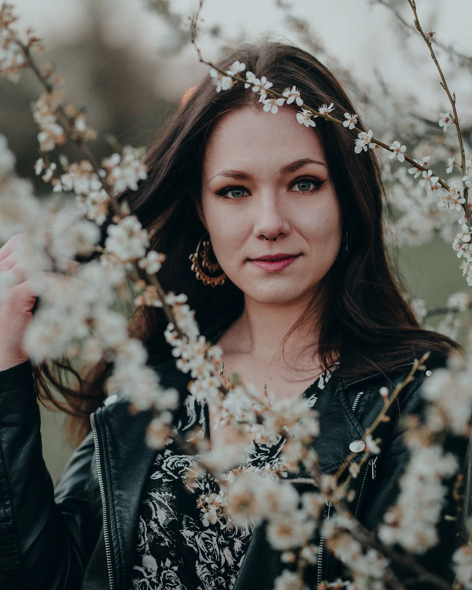 Porträt einer jungen Frau mit dunklem Haar, tätowierten Armen, Lederjacke und Ohrringen, die Blumen umrahmen.