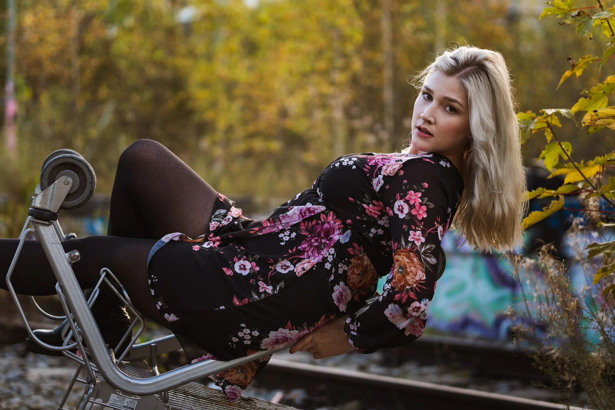 Junge Frau liegt auf einem Einkaufswagen vor einem Graffiti und Bäumen im Herbst.