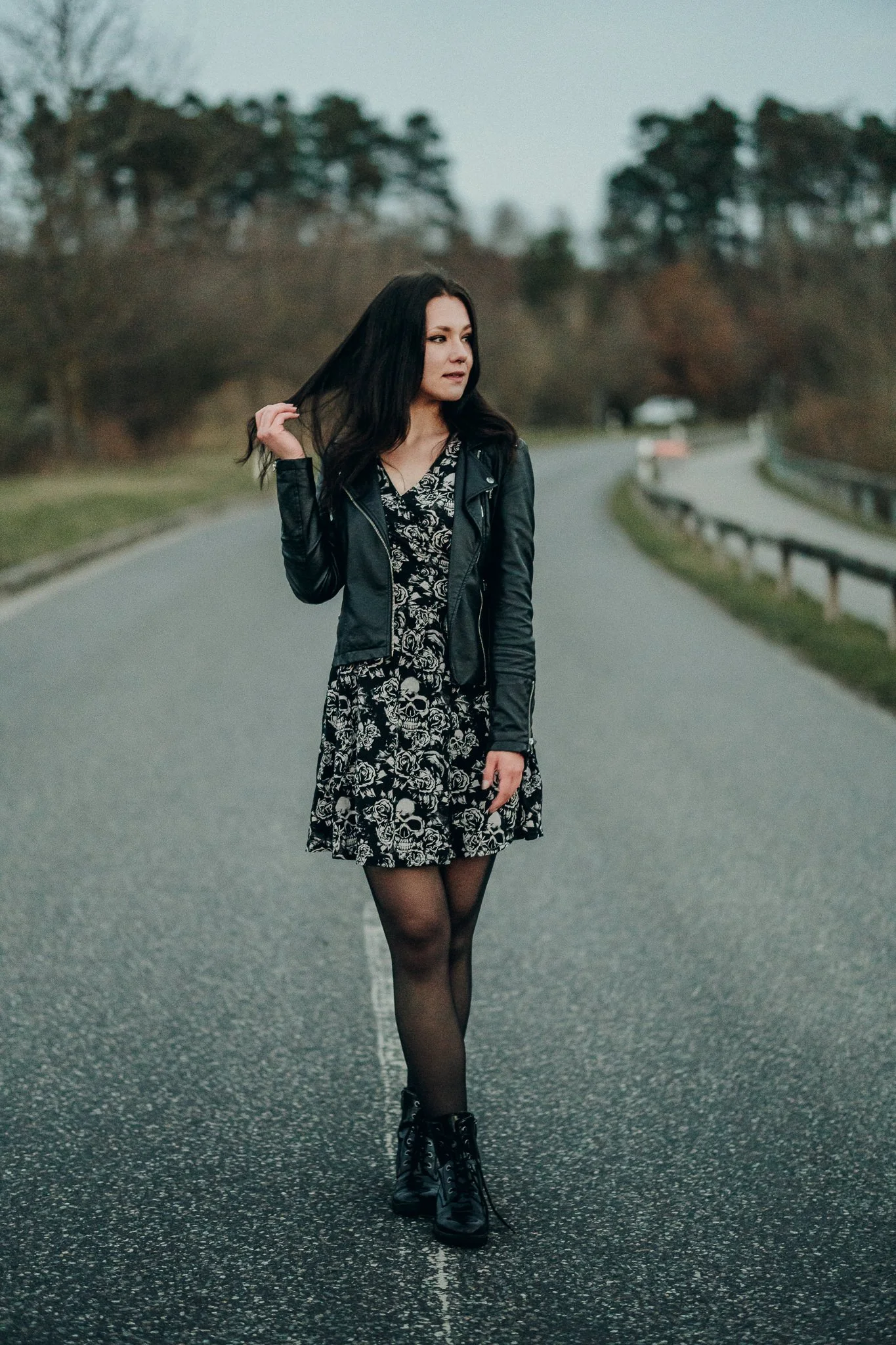 Junge Frau mit schwarzen Haaren trägt schwarze Jacke, floral-helles Kleid, schwarze Tights und Stiefel auf leere Straße im Freien.