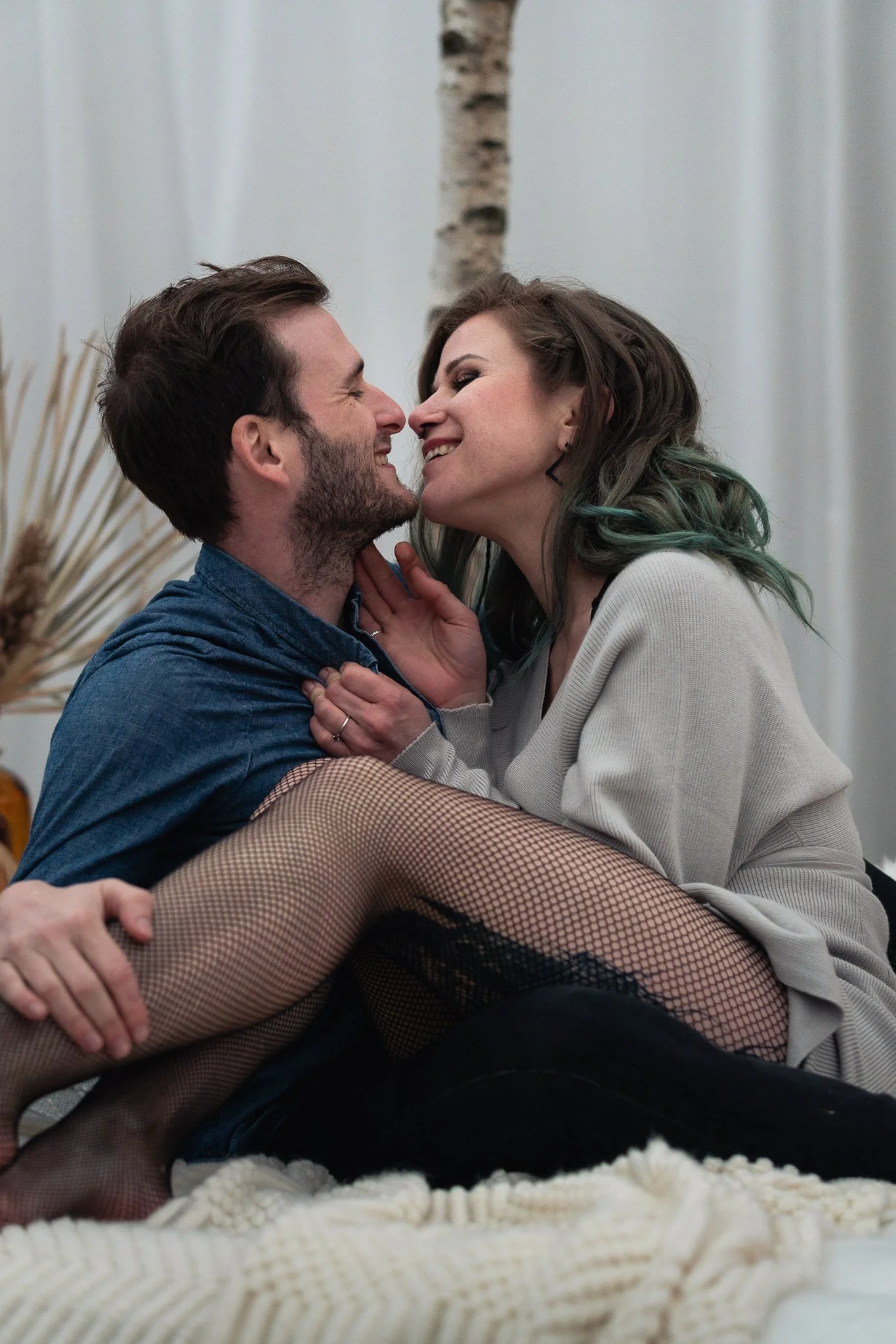 Paar in einer intimen Umarmung, lachen sich an, sitzen auf einem Bett, im Hintergrund ein Baumzweig und Deko.