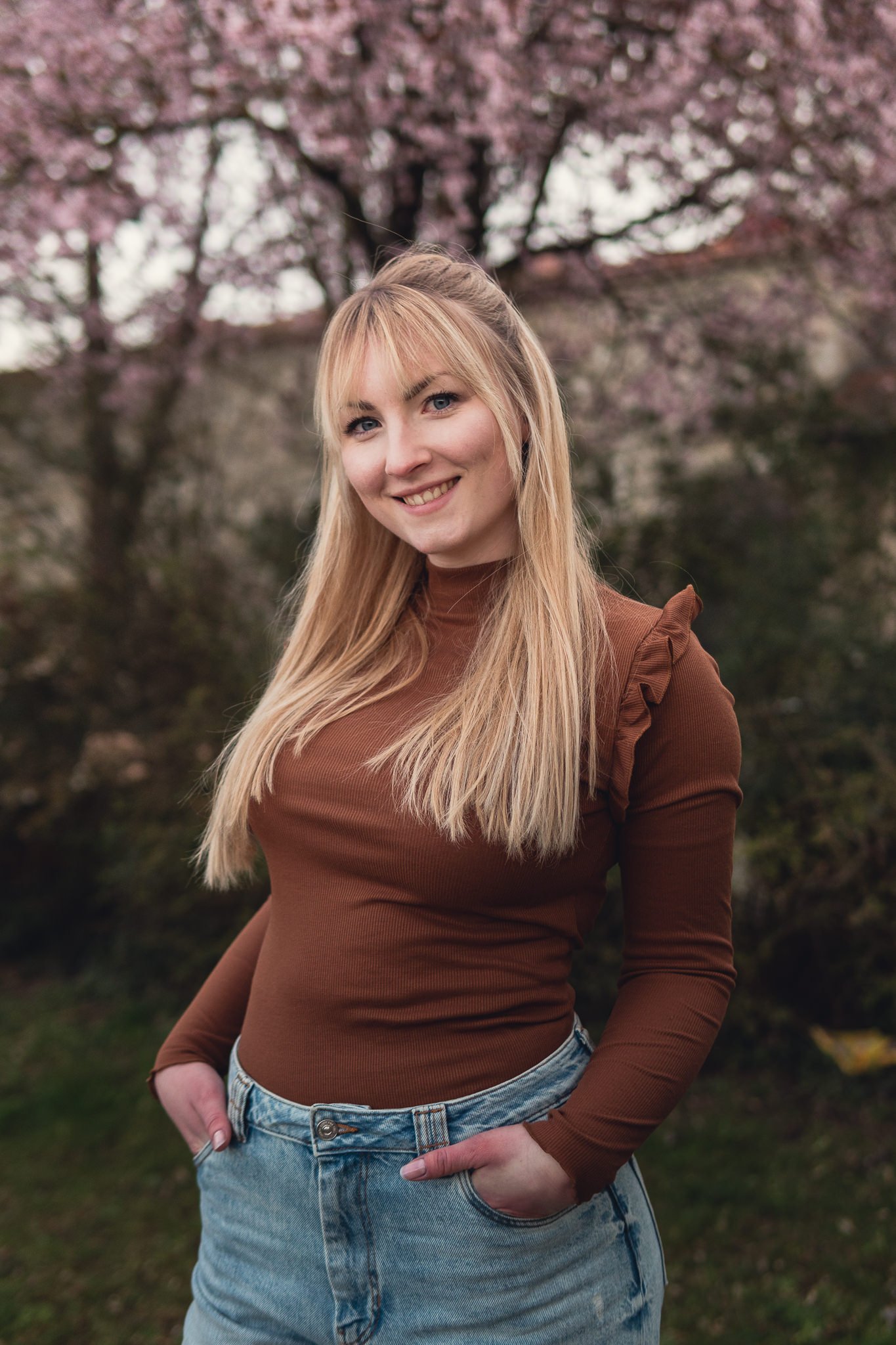 Eine lächelnde blonde Frau steht vor einem blühenden Baum mit rosa Blüten im Hintergrund.