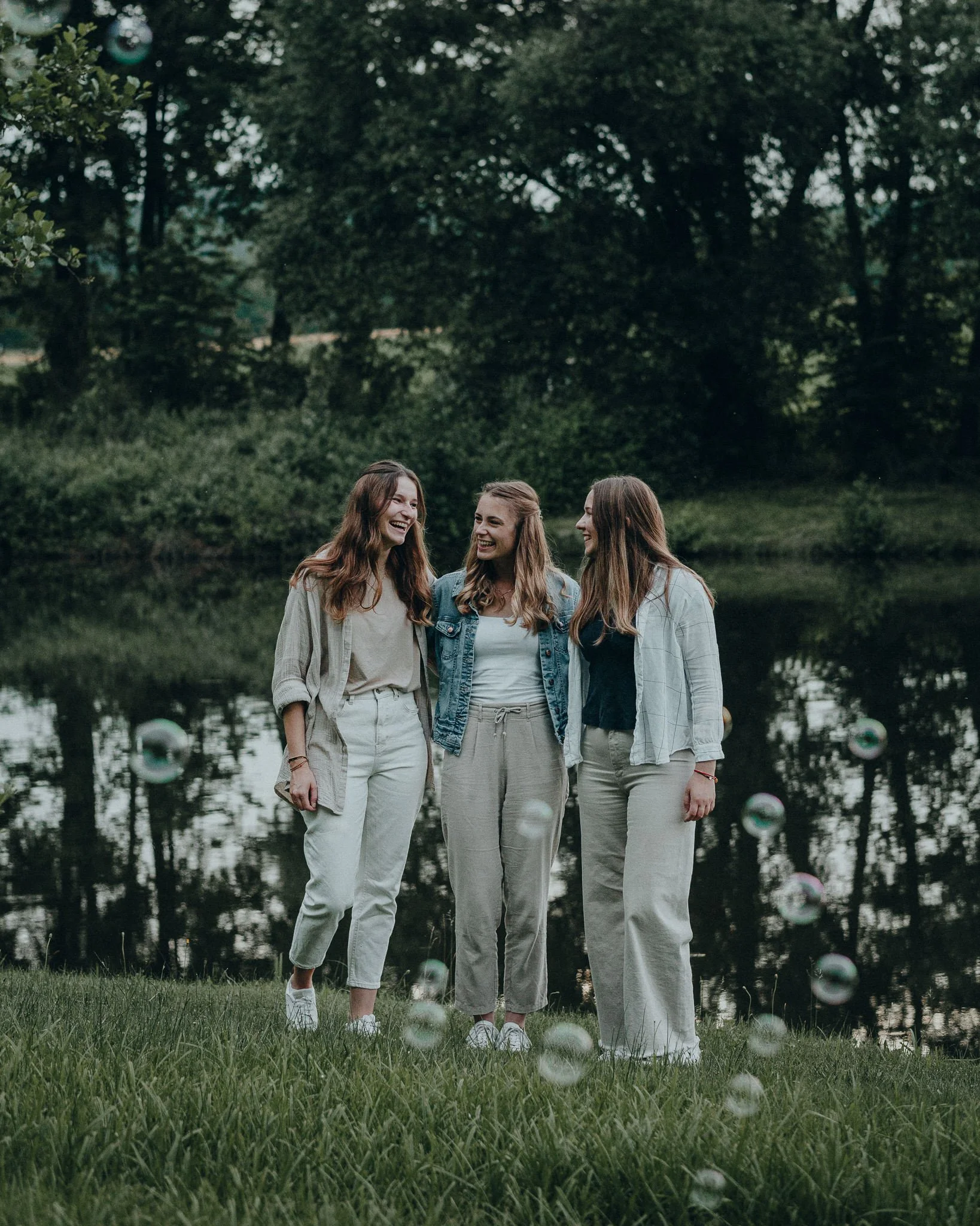 Drei lachende Frauen stehen am Ufer eines Sees im Grünen, mit Seifenblasen in der Luft, im Hintergrund Bäume.