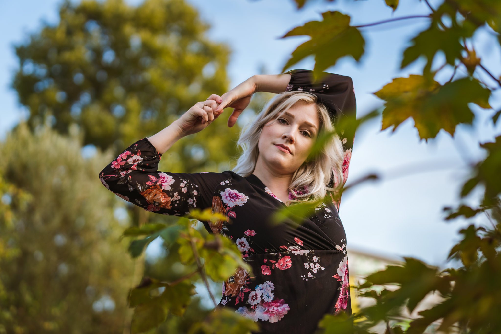 Junge Frau mit blonden, lockigen Haaren trägt ein schwarzes, lange Arm Kleid mit rosa und orange Blumen, steht im Grünen unter Bäumen, blickt in die Kamera, Arme über dem Kopf verschränkt.