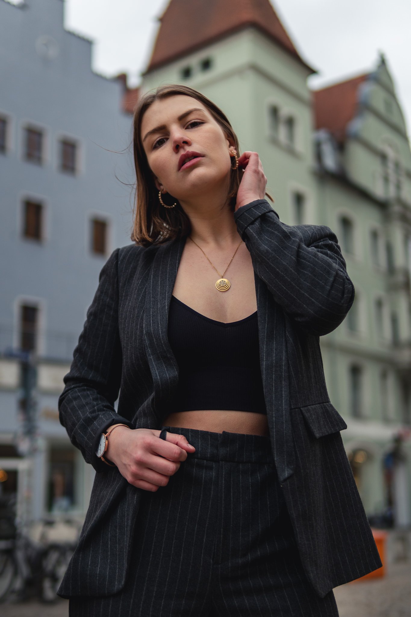 Junge Frau trägt einen schwarzen Business-Anzug mit Streifen, eine schwarze Top, Goldschmuck und eine Armbanduhr, steht in einer Stadt mit historischen Gebäuden im Hintergrund.