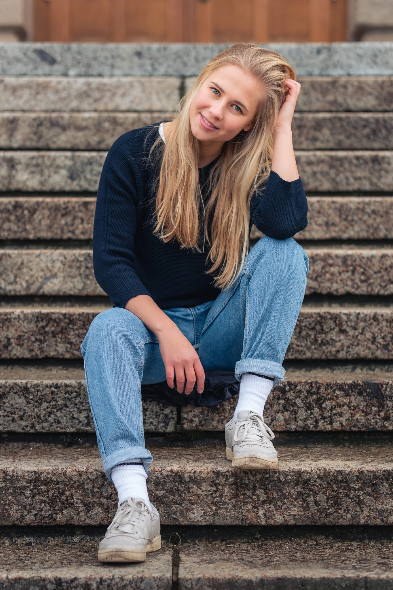 Junge Frau mit langen blonden Haaren, schwarzem Pullover, blauer Jeans, weißen Socken und Turnschuhen sitzt auf Steinstufen und lächelt in die Kamera.