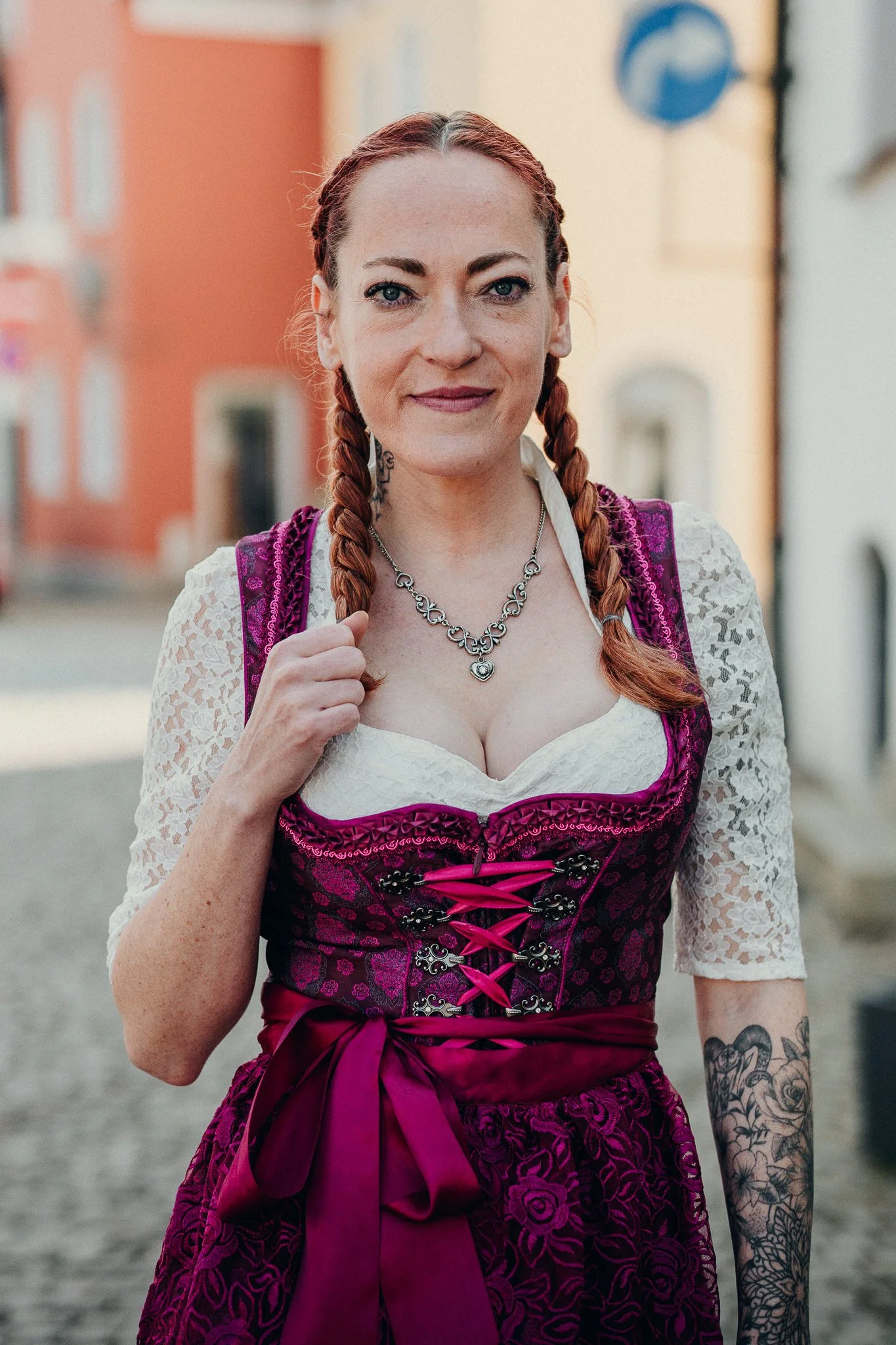 Frau in traditionellem bayerischem Dirndl mit roter Haarfrisur und Tätowierungen an den Armen, steht auf einer Straße mit alten Gebäuden im Hintergrund.