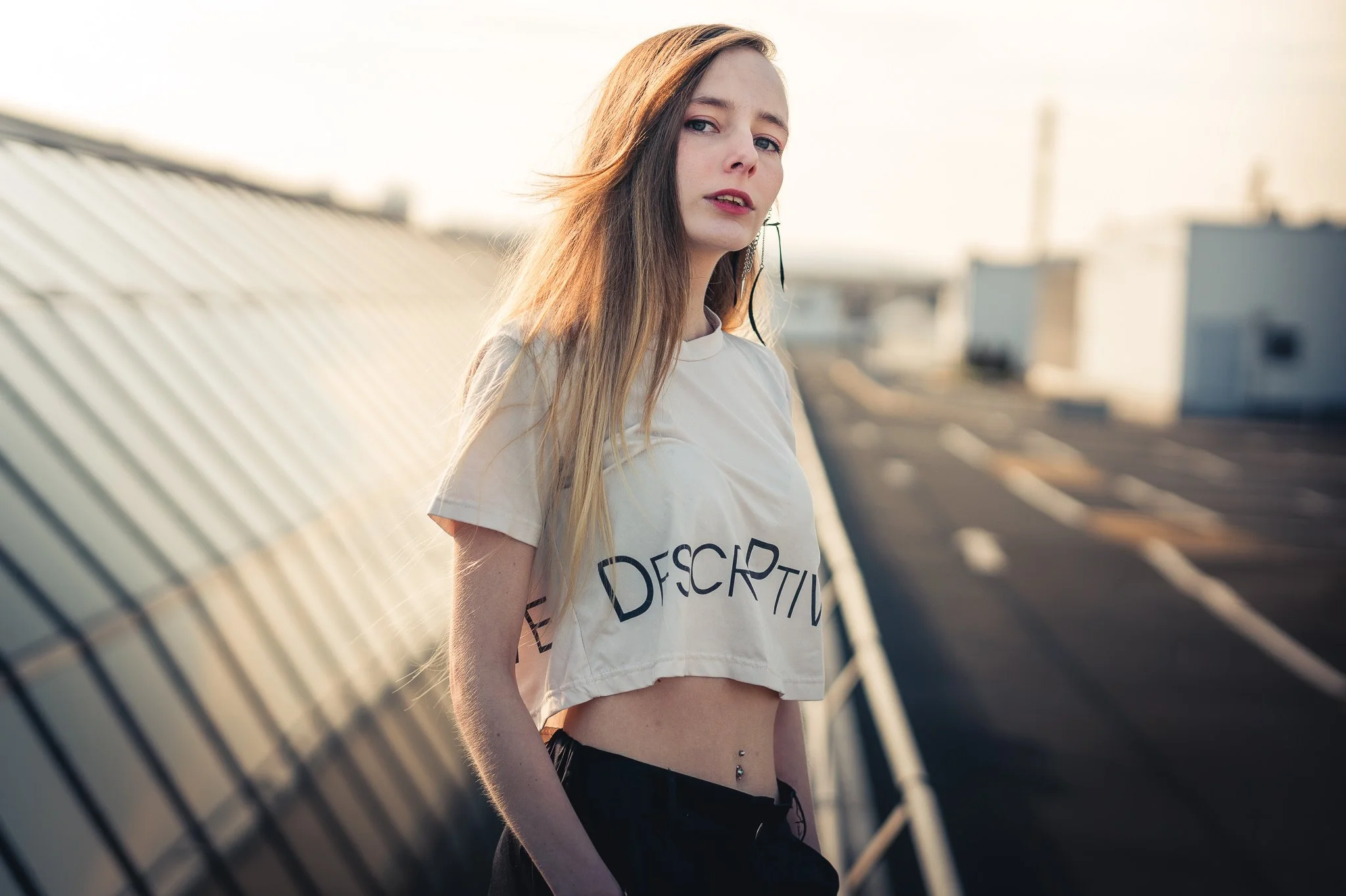 Junge Frau mit langem Haar, trägt ein weißes T-Shirt mit schwarzem Text, steht auf einem Dach bei Sonnenuntergang.