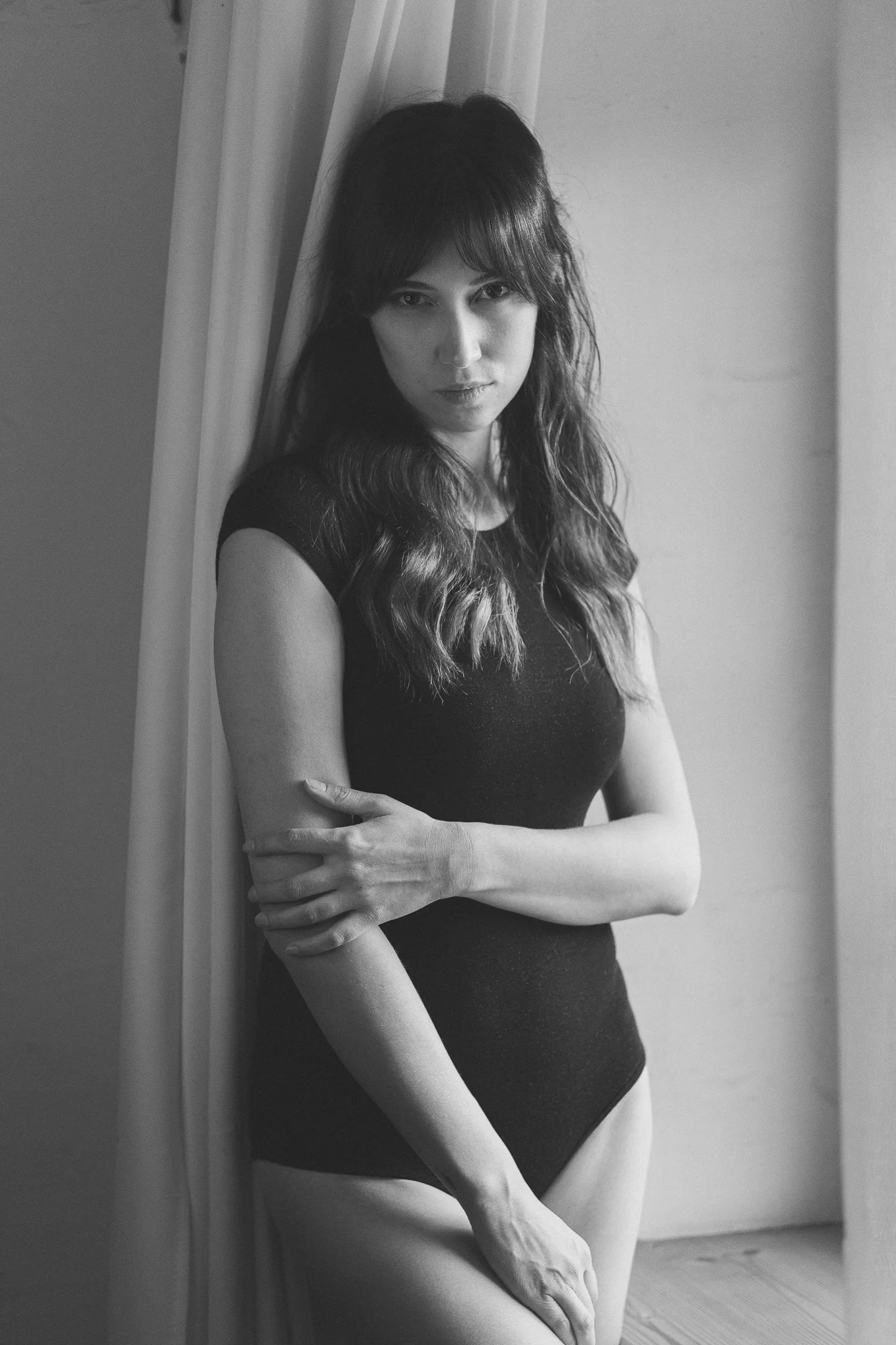 Schwarz-weiß-Fotografie einer jungen Frau in dunklem Kleid, die an einer Tür lehnt, mit langem, gewelltem Haar und einem ernsten Blick, leicht den Kopf geneigt.