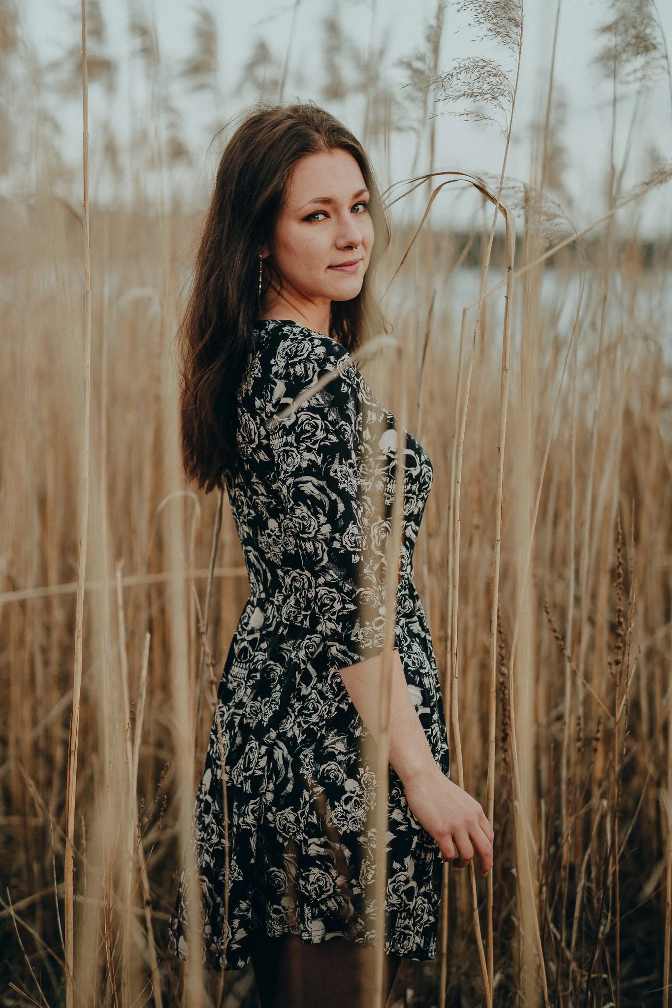 Eine Frau steht in einem Feld mit hohen Gräsern, trägt ein schwarzes Kleid mit weißen Rosenmustern, blickt zur Kamera und lächelt leicht.