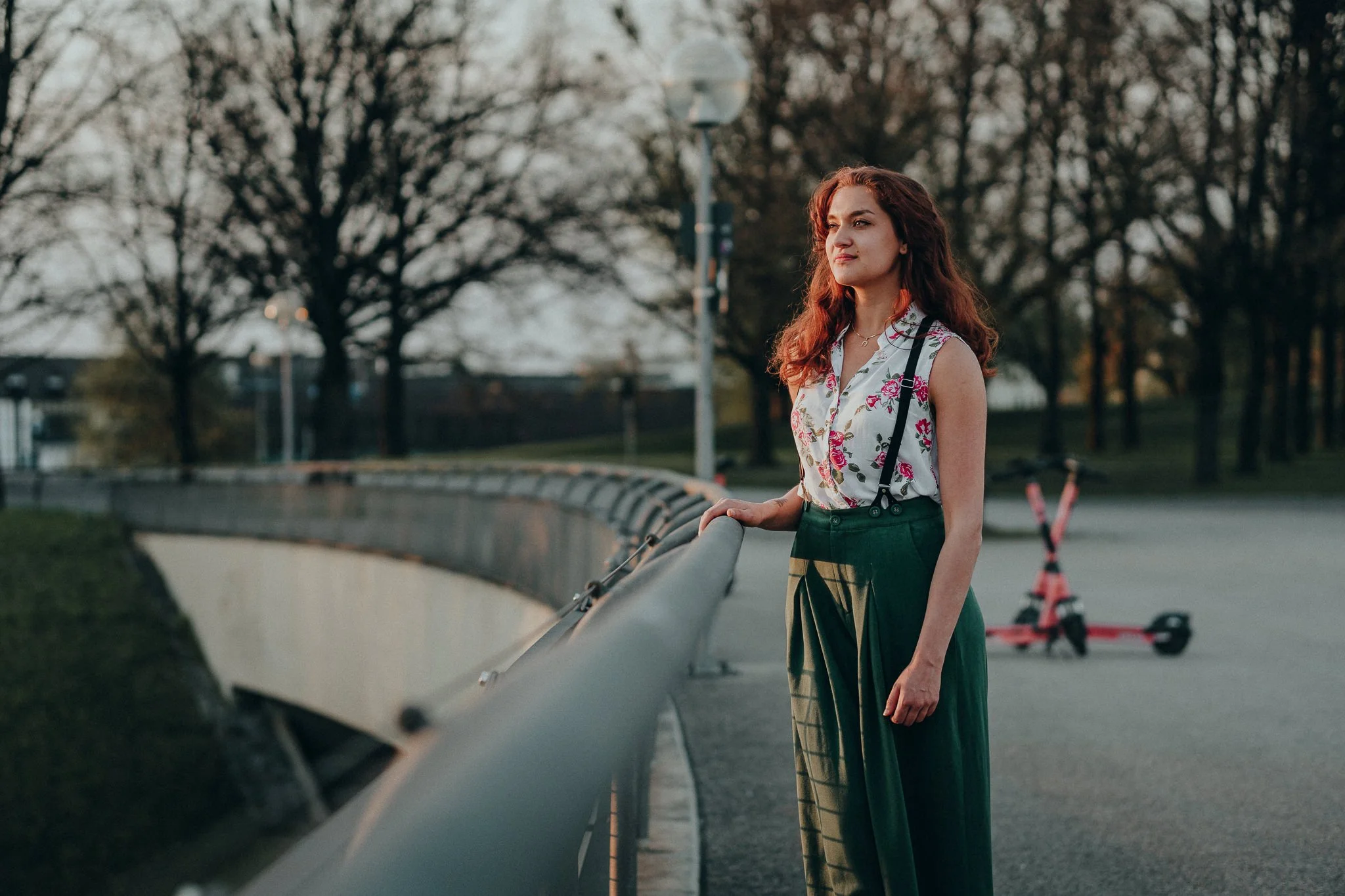 Eine junge Frau mit roten, lockigen Haaren steht an der Straßenbarriere und schaut in die Ferne. Sie trägt ein weißes ärmelloses Oberteil mit Blumenmuster und dunkle, weite Hosen. Im Hintergrund sind Bäume und elektrische Scooter sichtbar, die Szener