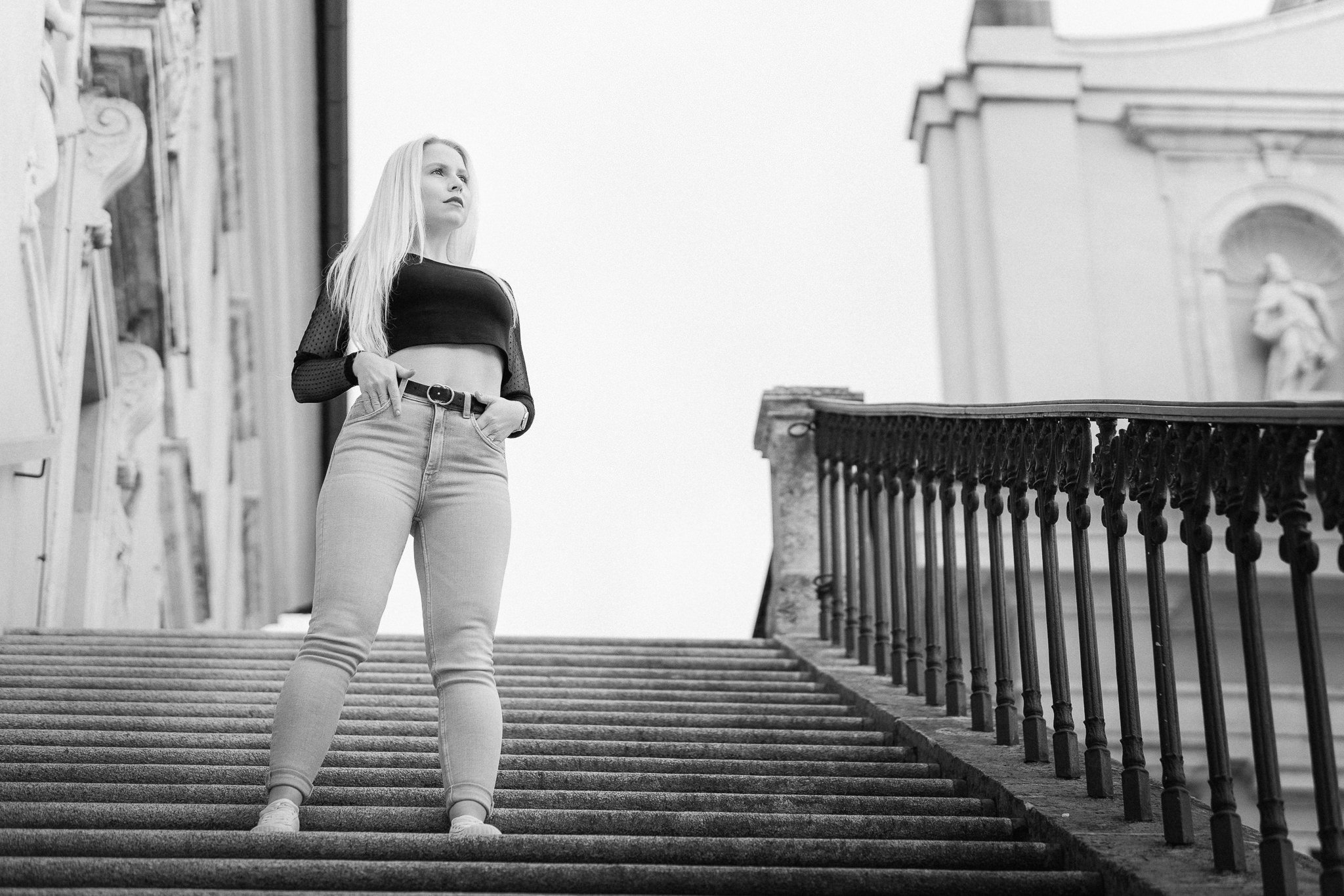 Junge Frau mit langen Haaren steht auf Steintreppe vor historischen Gebäuden, blickt nach oben, trägt schwarze Top, gepunktete Überwurfjacke, Jeans und Sneakers, schwarz-weiß Foto