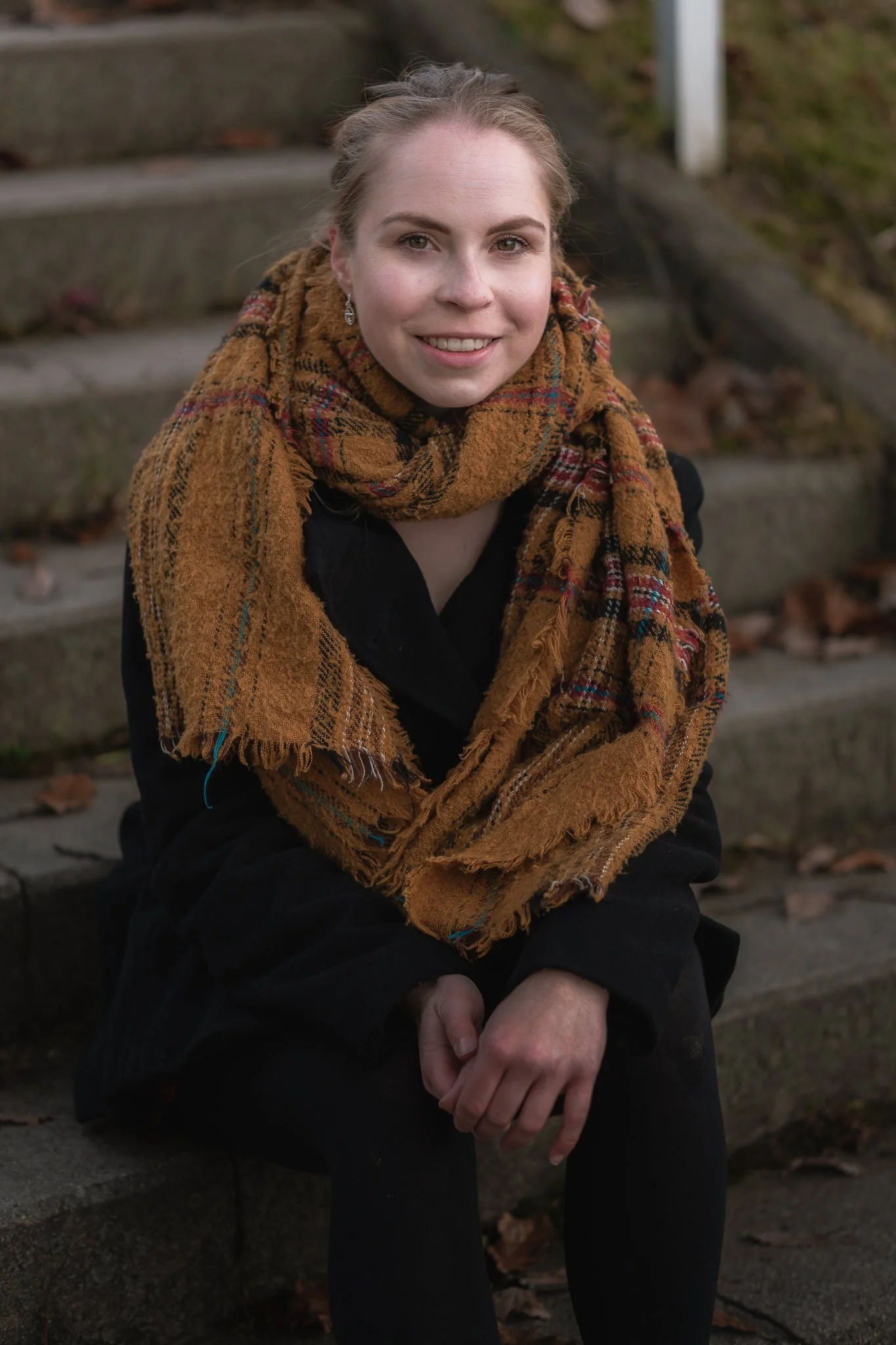 Junge Frau mit braunem Schal und schwarzer Jacke sitzt auf Stein-Stufen im Freien, im Herbst, lächelnd.