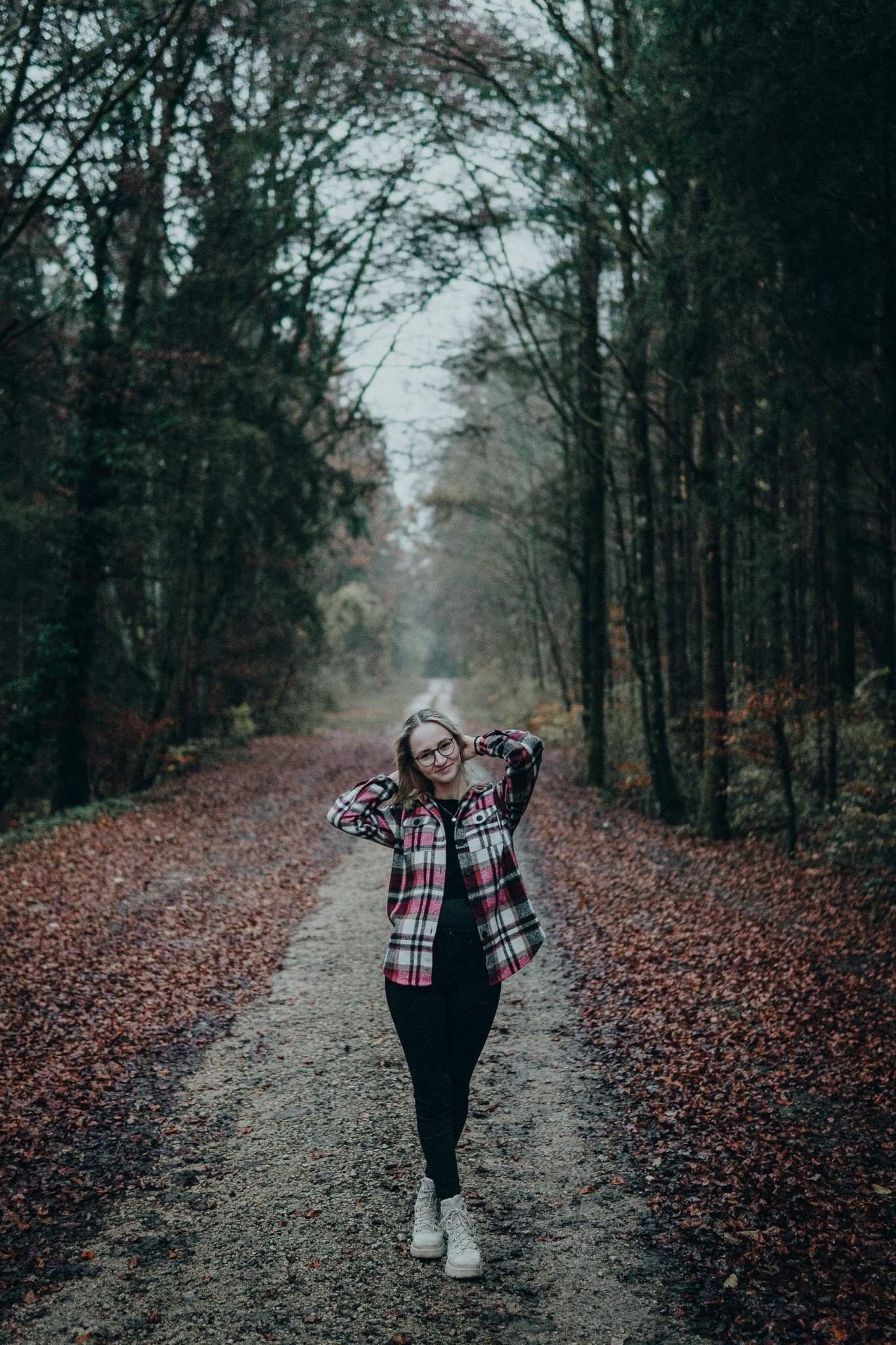 Eine jungfrau Frau trägt eine rote, weiße und schwarze karierte Jacke, schwarze Hose und weiße Schuhe, steht auf einem Waldweg im Herbst, umgeben von Bäumen mit fallendem Laub.