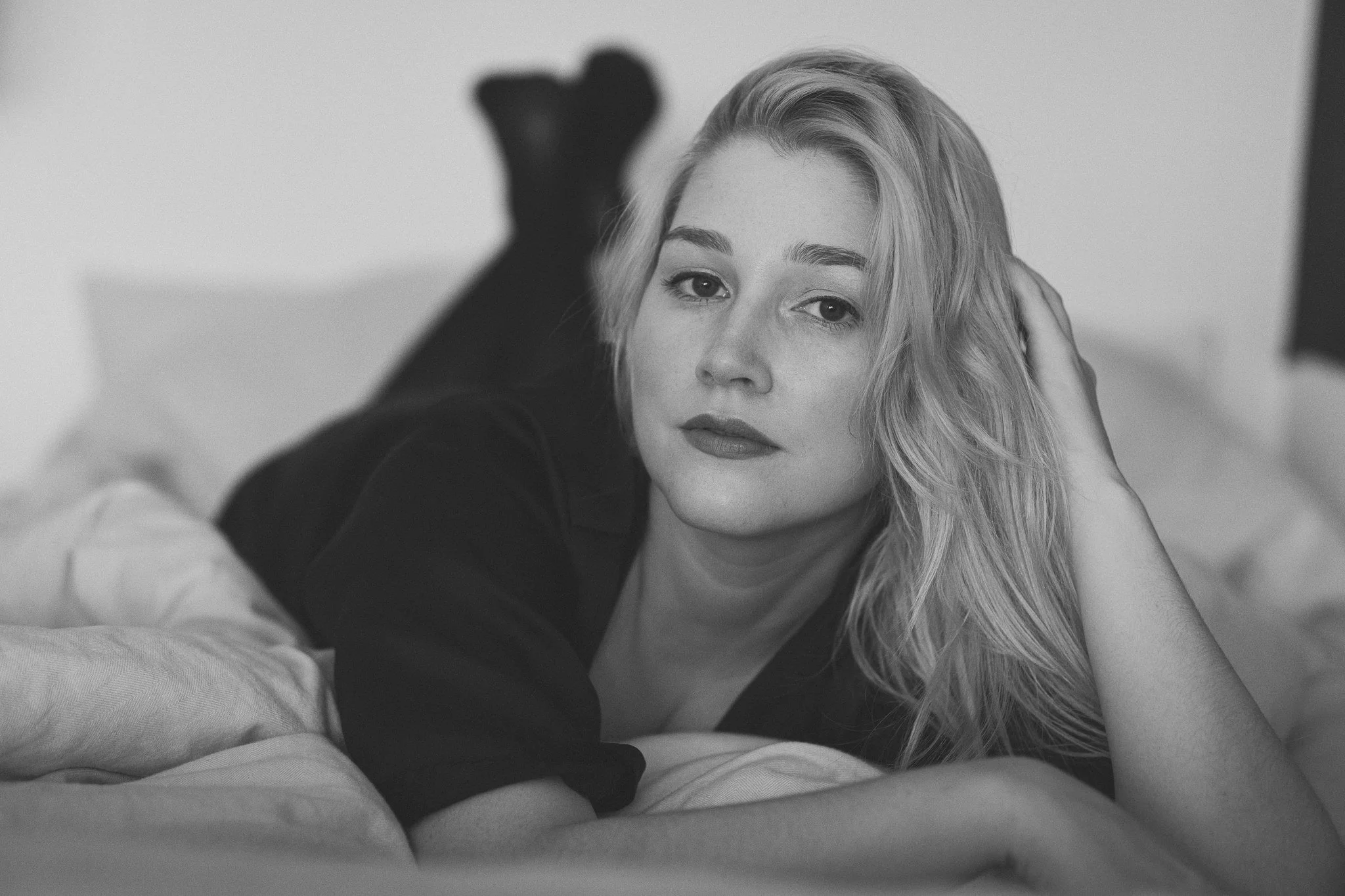 Junge Frau liegt auf einem Bett, ruht ihren Kopf auf ihrer Hand und blickt in die Kamera, im Hintergrund unscharfe Wand.